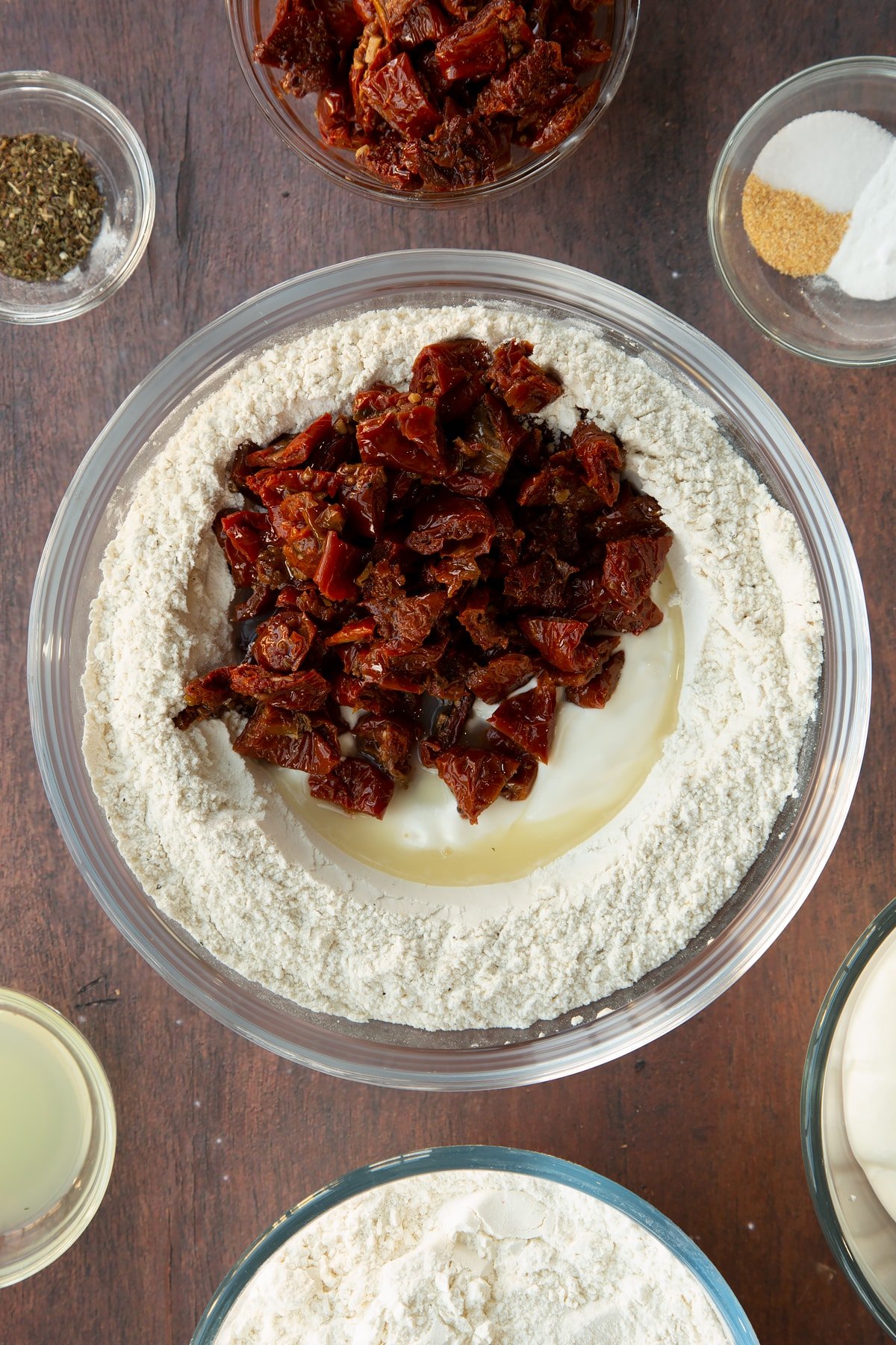 Flour, bicarbonate of soda and spices in a bowl with a well in the middle filled with yogurt, lemon juice and sundried tomatoes. Ingredients to make Sundried tomato soda bread surround the bowl.