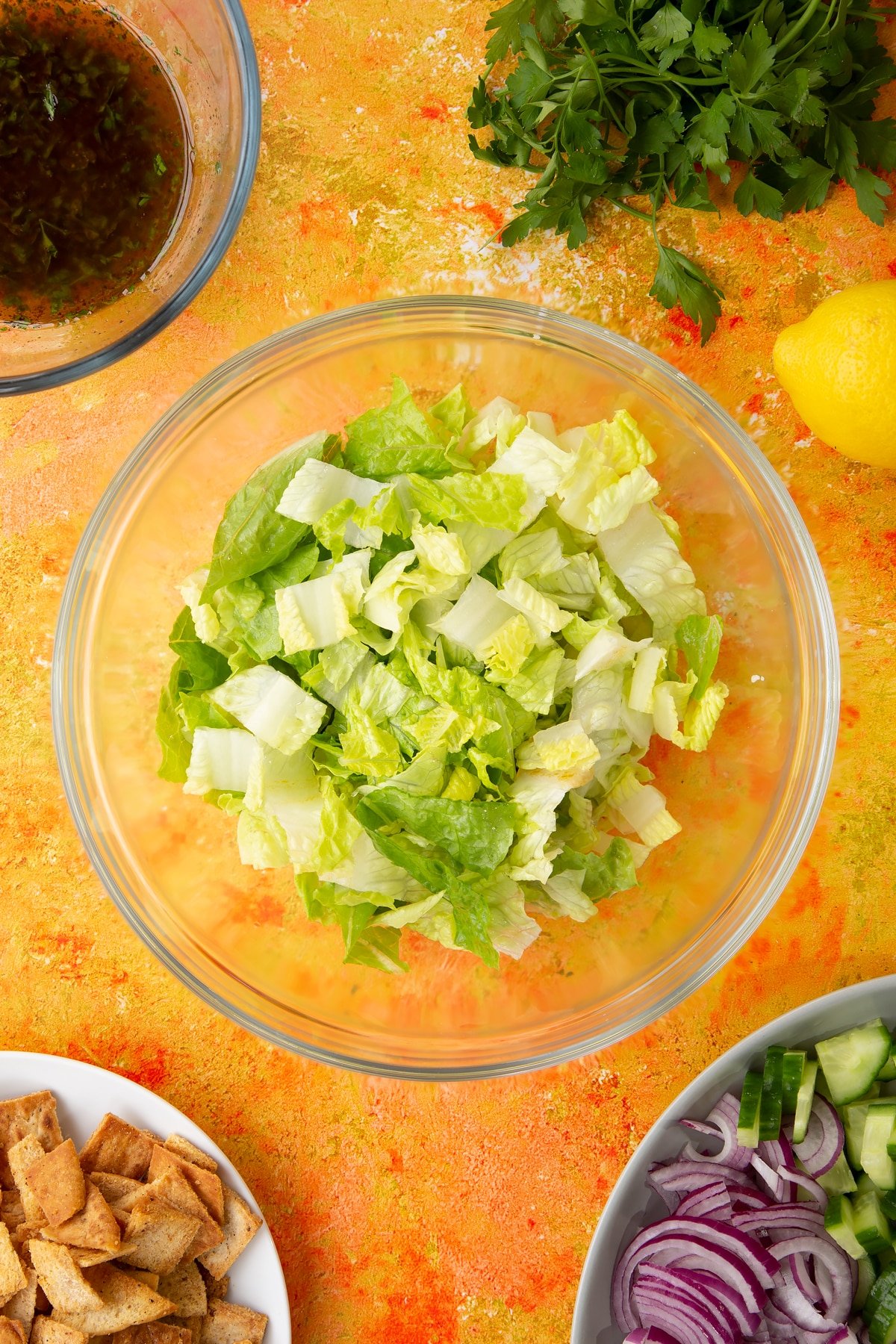 Lettuce in a glass mixing bowl. Ingredients to make vegan fattoush surround the bowl.