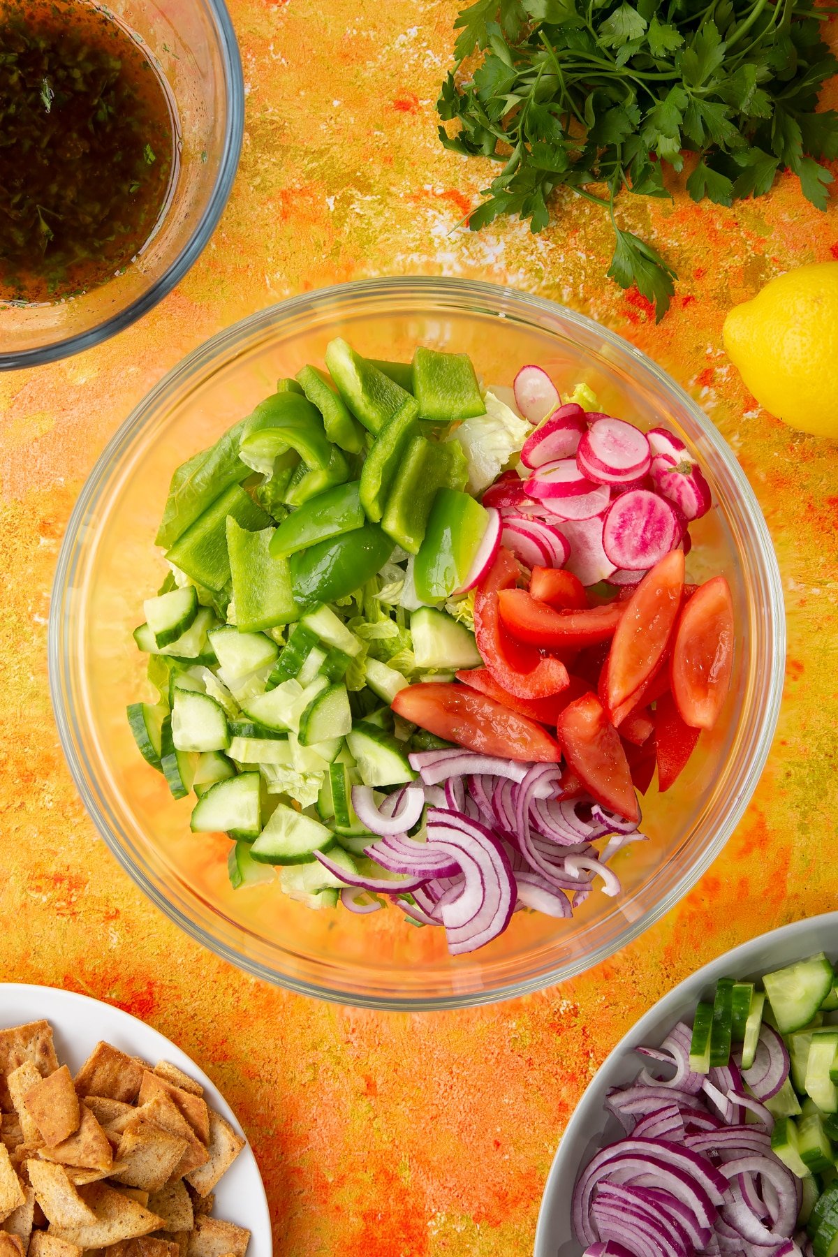 Lettuce, tomatoes, red onion, cucumber, green peppers and radish in a glass mixing bowl. Ingredients to make vegan fattoush surround the bowl.
