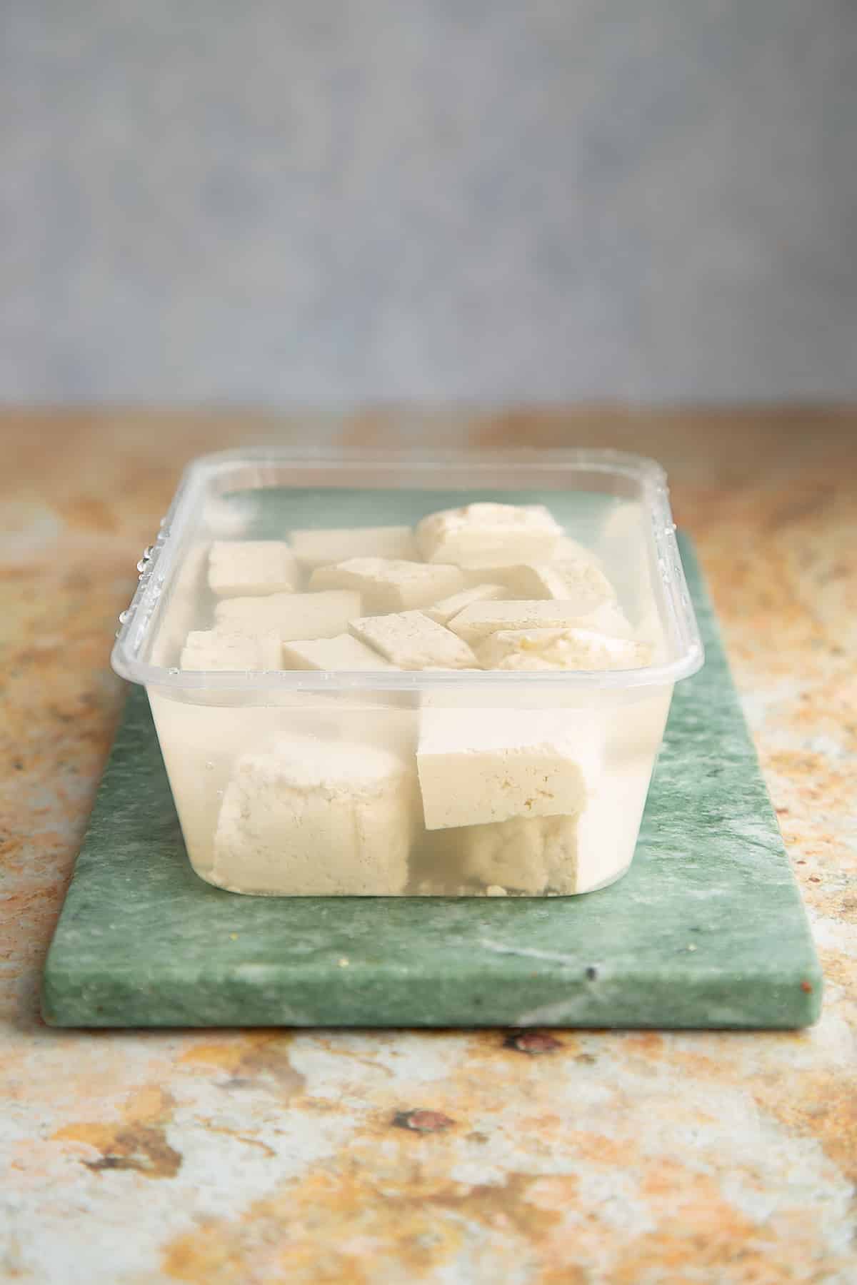 cubed tofu in a plastic container covered in brine sat on a green marble board.