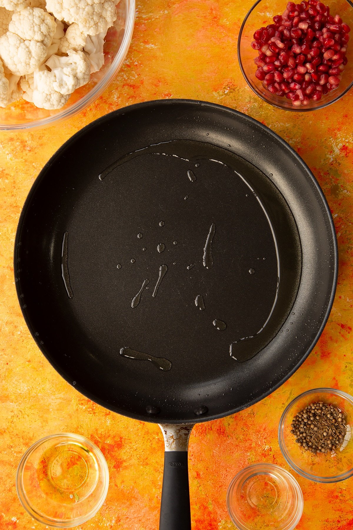 Overhead shot of oil in a pan with ingredients for the cauliflower couscous with pomegranate surrounding it. 