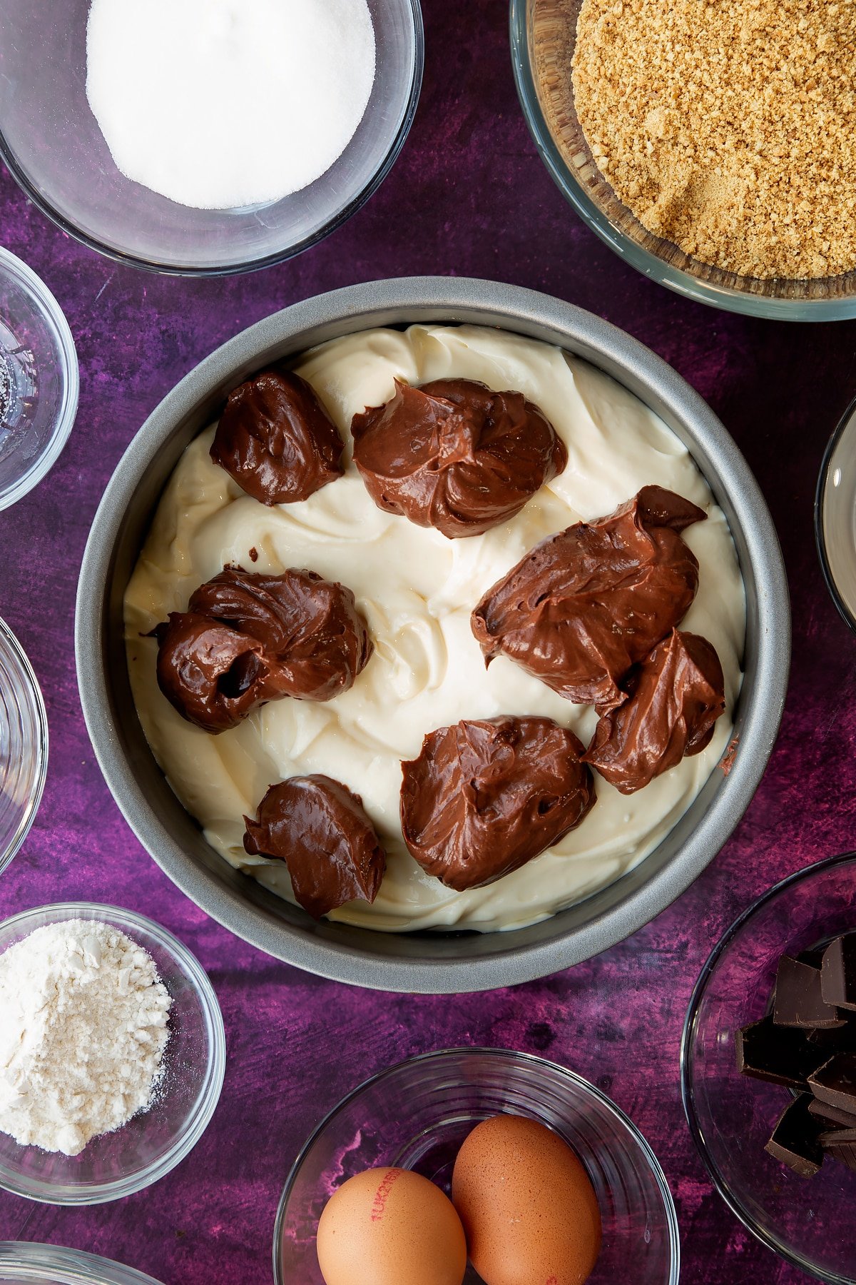 Adding spoonfuls of the chocolate cream cheese filling for the marble cheesecake to the non-stick cake tin.