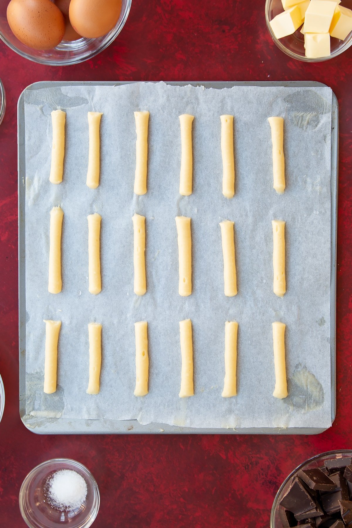 Thin lines of choux from the mini eclairs pastry piped directly onto the greaceproof paper atop the baking tray.