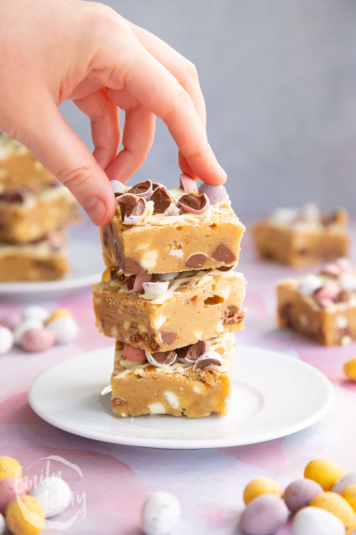Three Mini Egg blondies on a white plate. More Mini Egg blondies are behind. A hand reaches for the top one.