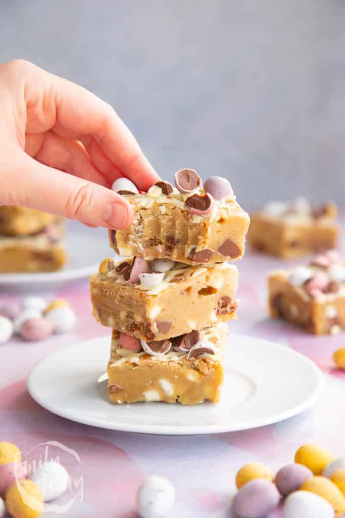 Three Mini Egg blondies on a white plate. More Mini Egg blondies are behind. The top one has been bitten and a hand reaches for it.