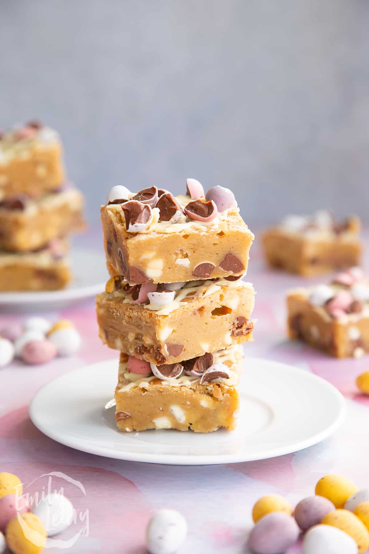 Three Mini Egg blondies on a white plate. More Mini Egg blondies are behind.