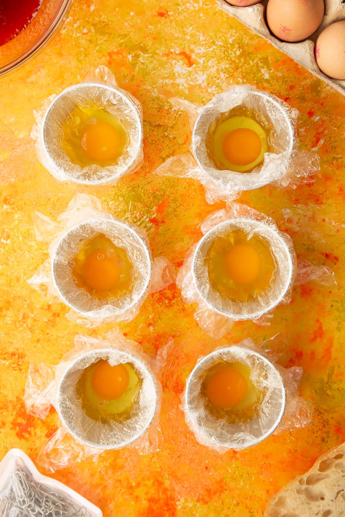 Overhead shot of six small bowls i.e. ramkins covered in clingfilm and oil with an egg cracked inside.