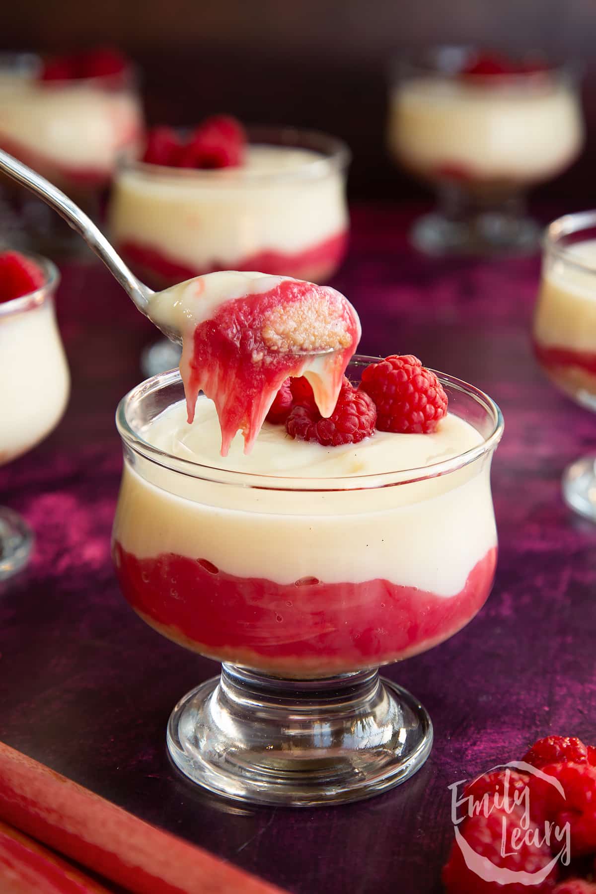 Rhubarb and custard pudding in a glass dish topped with raspberries with a spoonful on show.