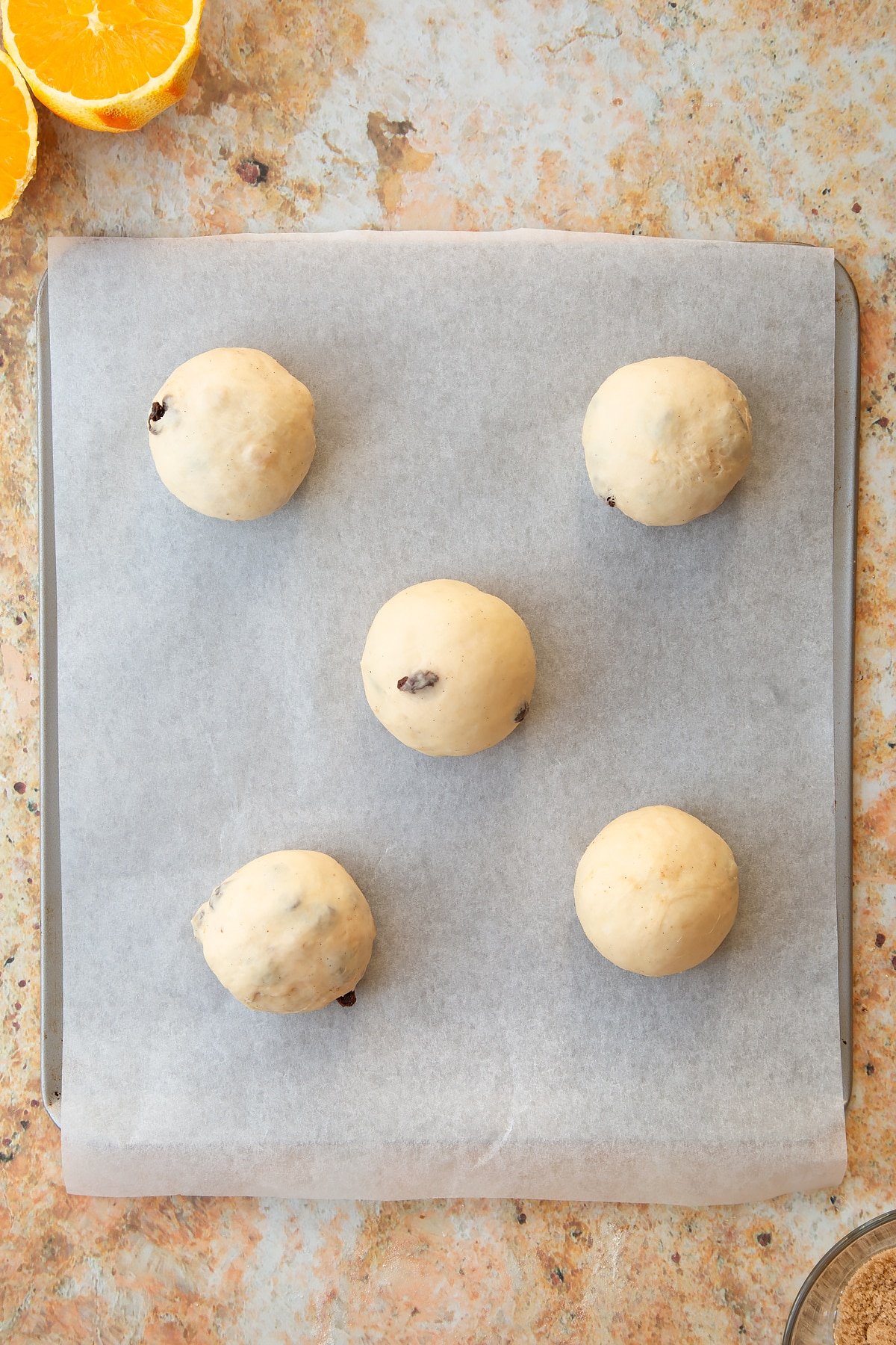 Overhead shot of the spiced fruit buns dough balls on a baking sheet ontop of a baking tray.