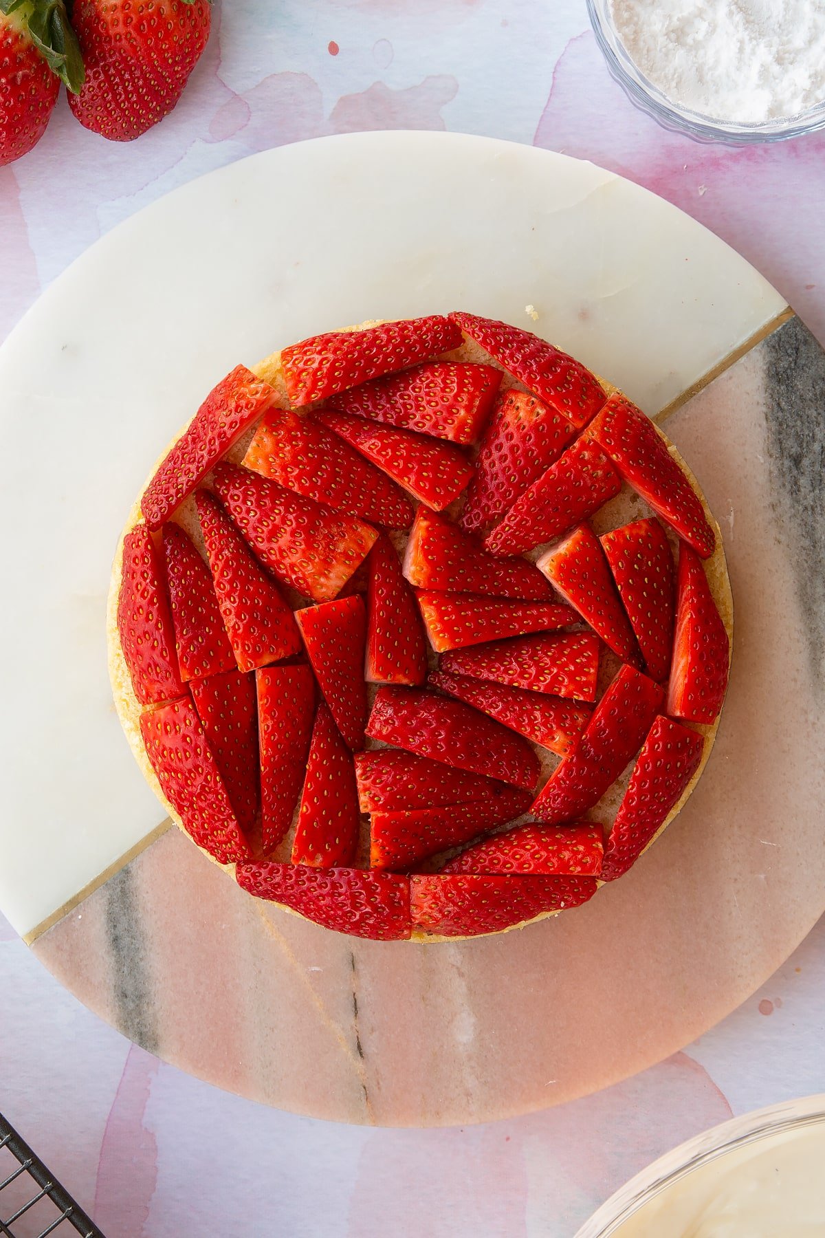 circular sponge cake on a pink marble  board topped with chopped strawberries.