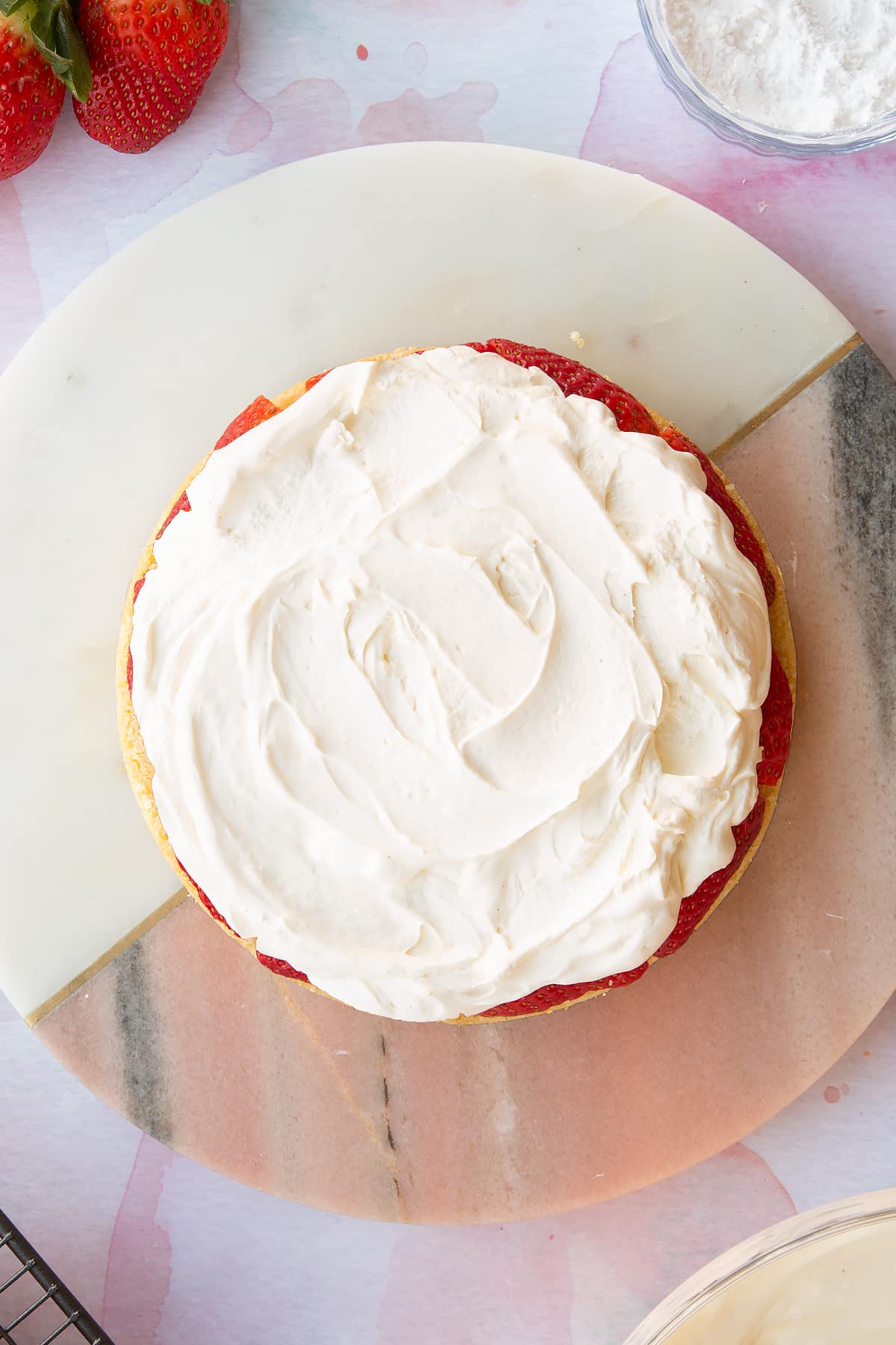 circular sponge cake topped with chopped strawberries and fresh cream on a pink marble  board.