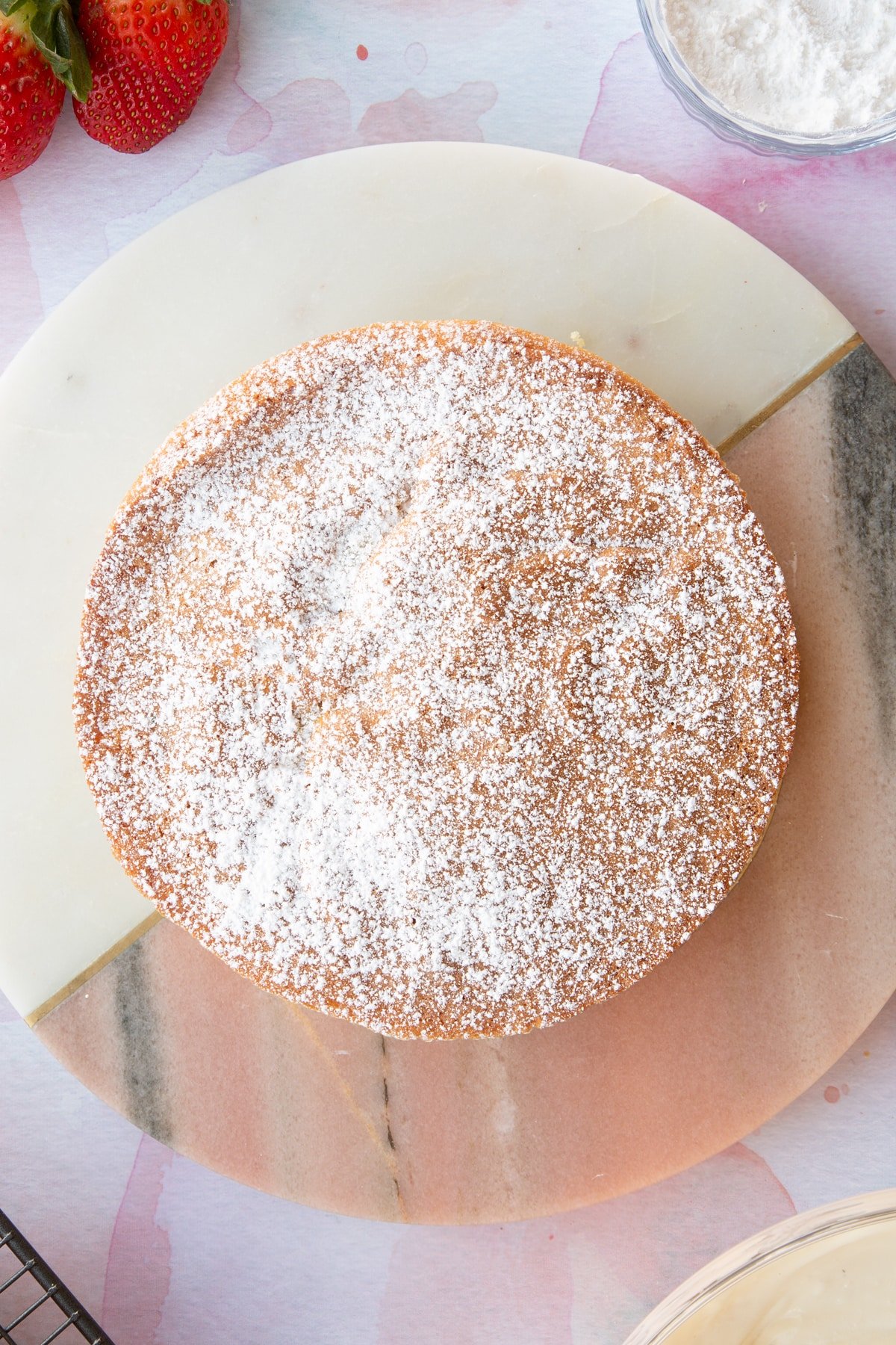 circular strawberry cream sponge cake topped with a dusting og icing sugar on a pink marble  board.