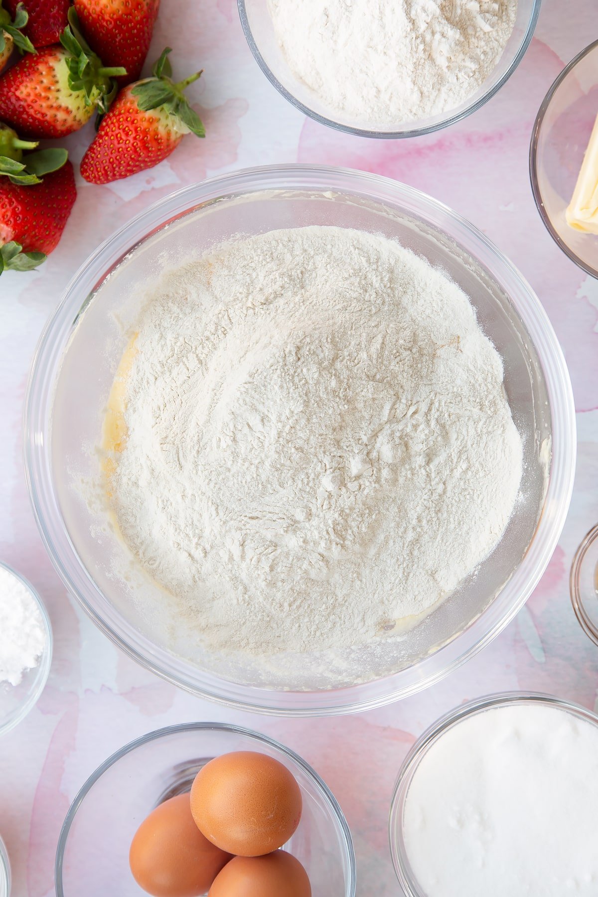 creamed butter, sugar and egg mixture topped with flour in a large clear bowl with ingredients around the edges 