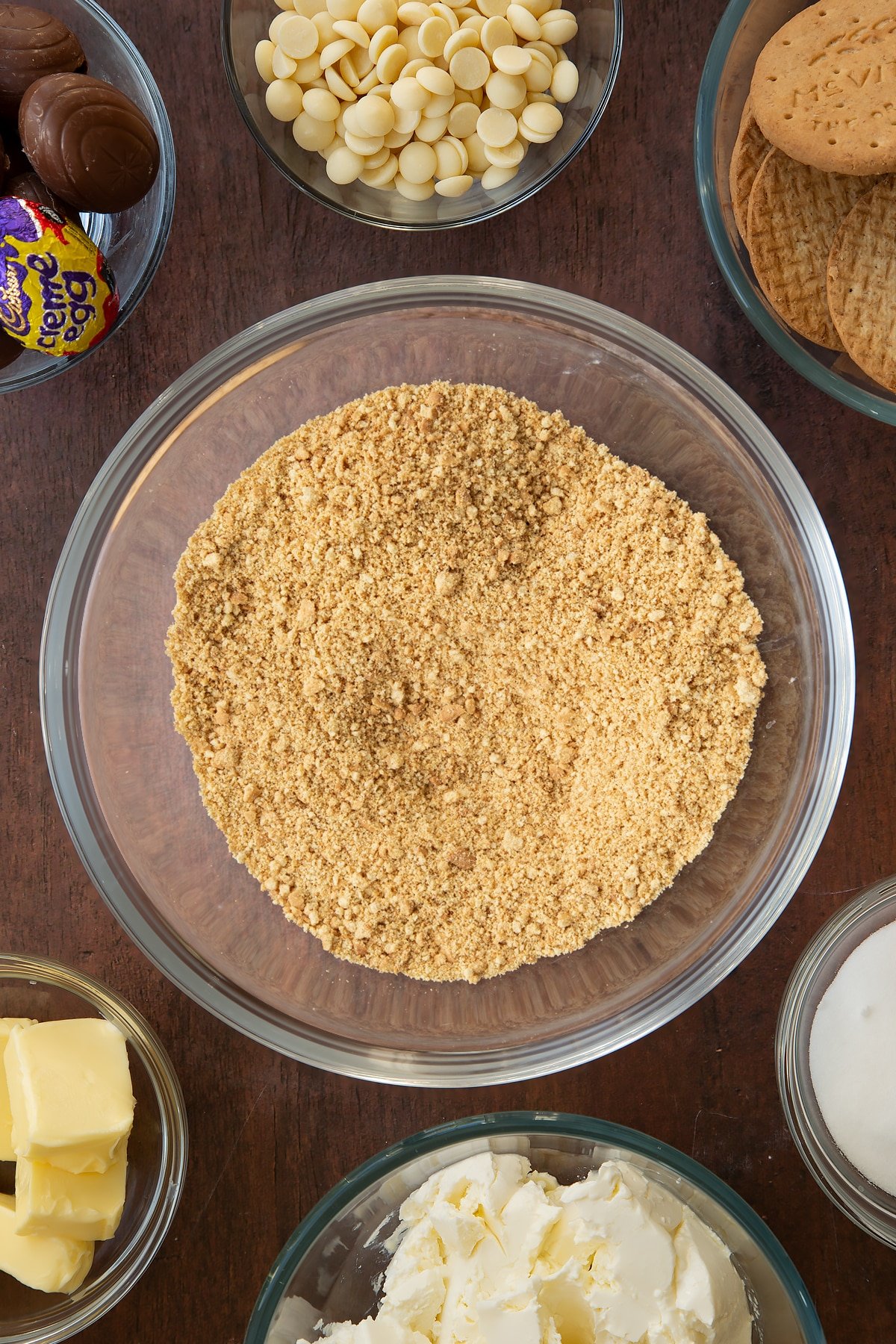 Crumbled biscuits in a mixing bowl with ingredients for the Cadbury Creme Egg cheesecake on the side.