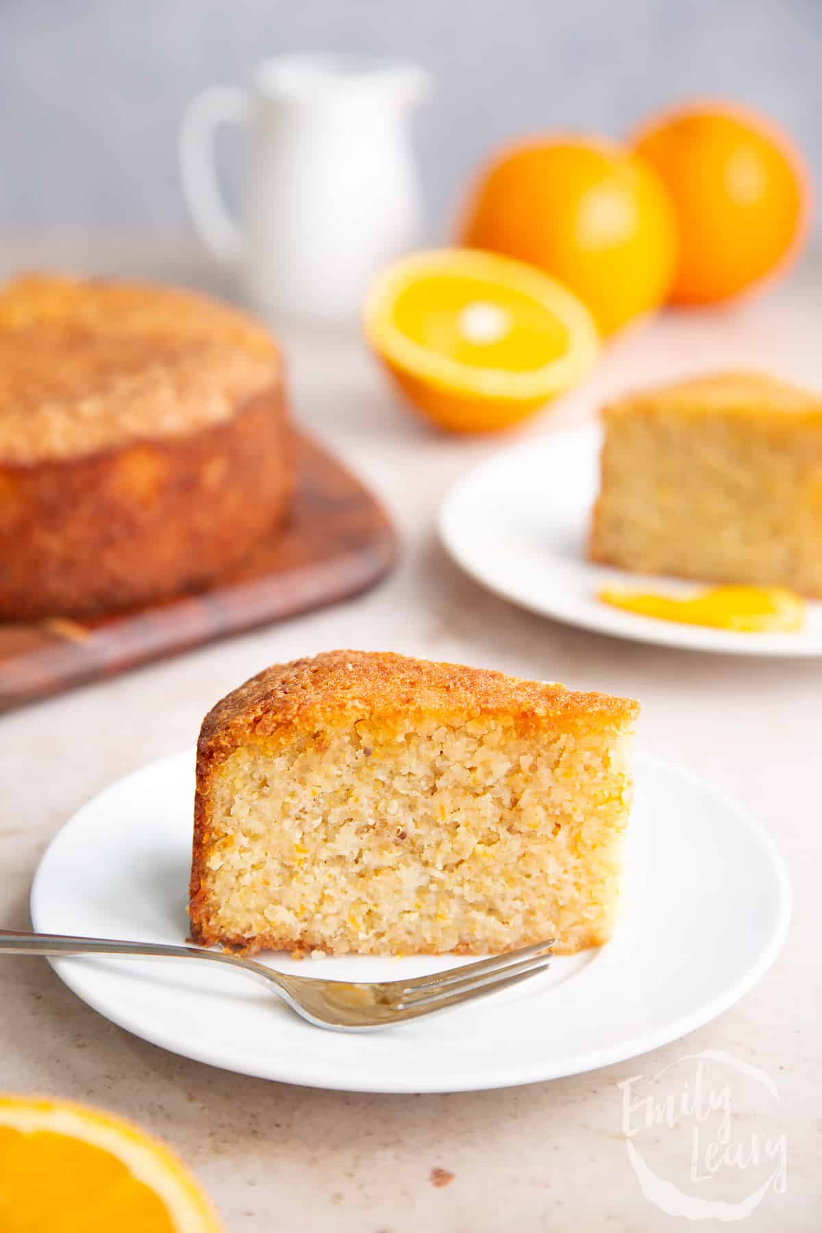 White plate with a small slice of gluten free orange cake ontop. A fork on the side and additional gluten free orange cake slices in the background.