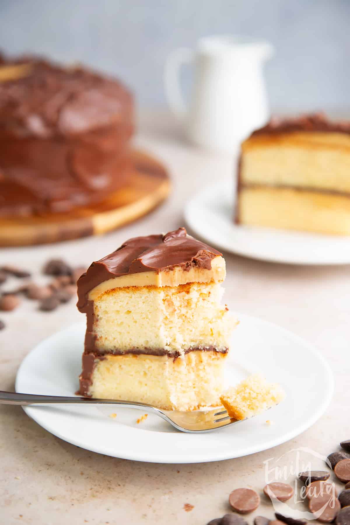 a slice of Millionaire shortbread cake on a white plate with  a fork cutting off the front point and the full cake in the background.