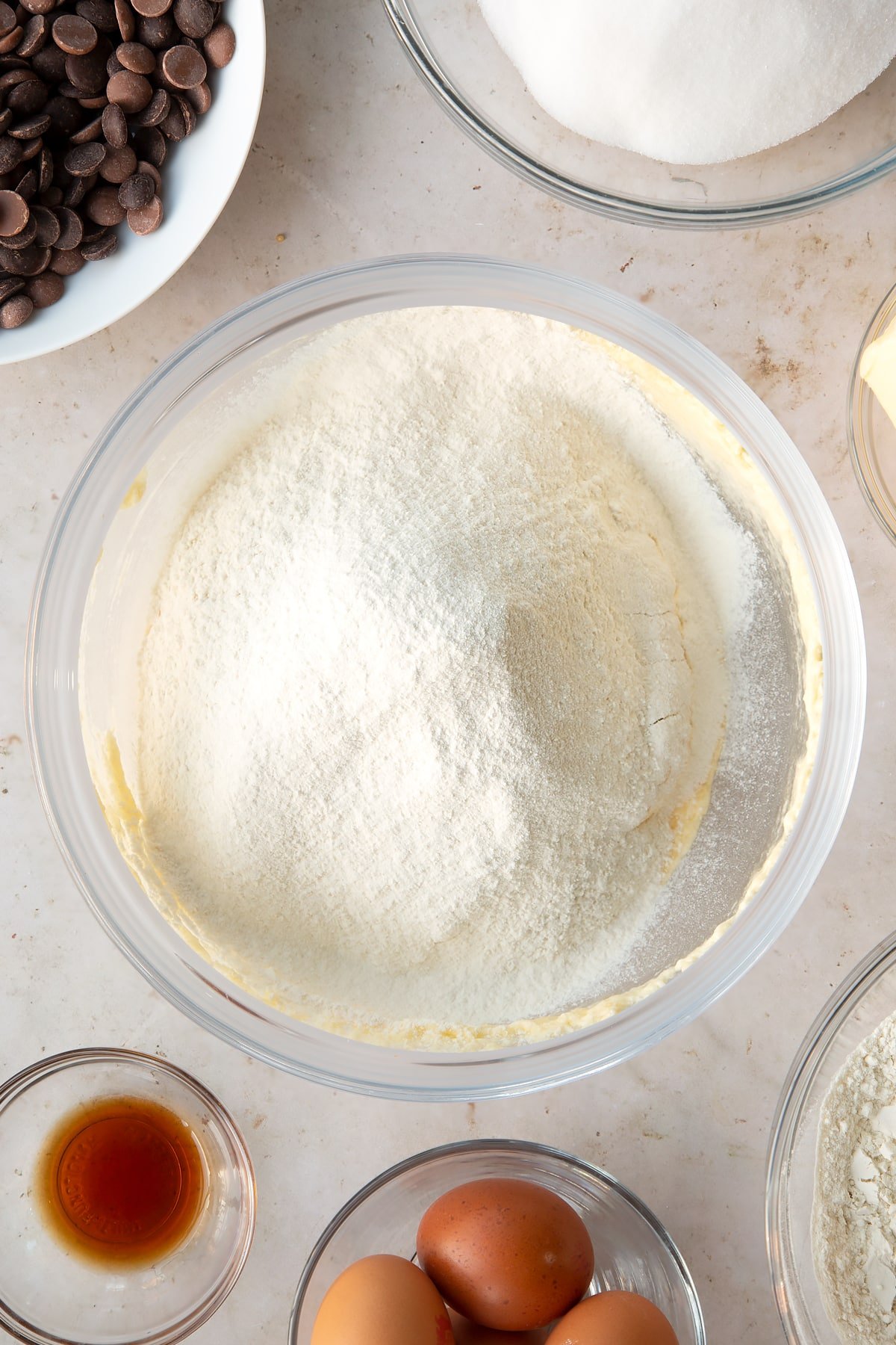 whisked cake batter in a large clear bowl topped with flour surrounded by ingredients.