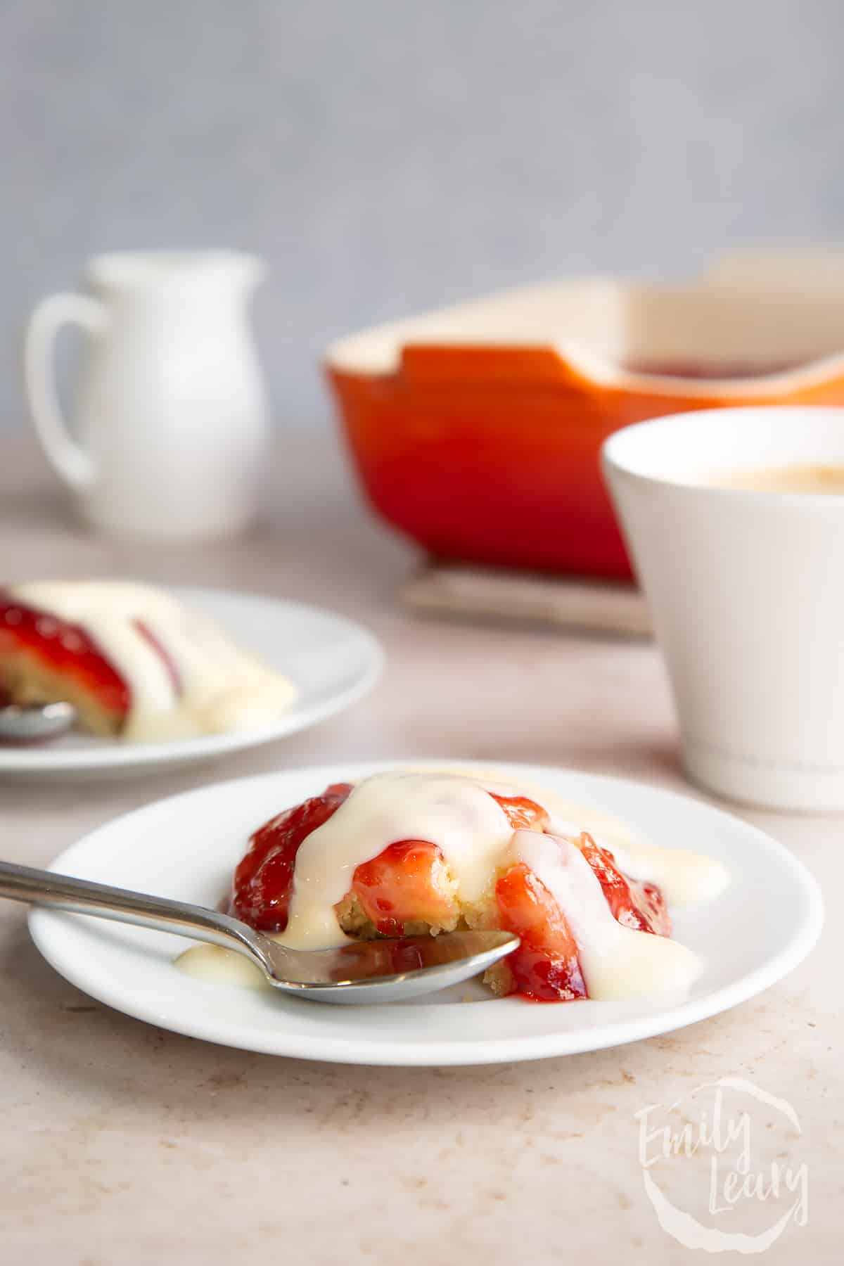 side view of Vegan jam sponge pudding on a white plate with a spoon on the side and topped with custard.