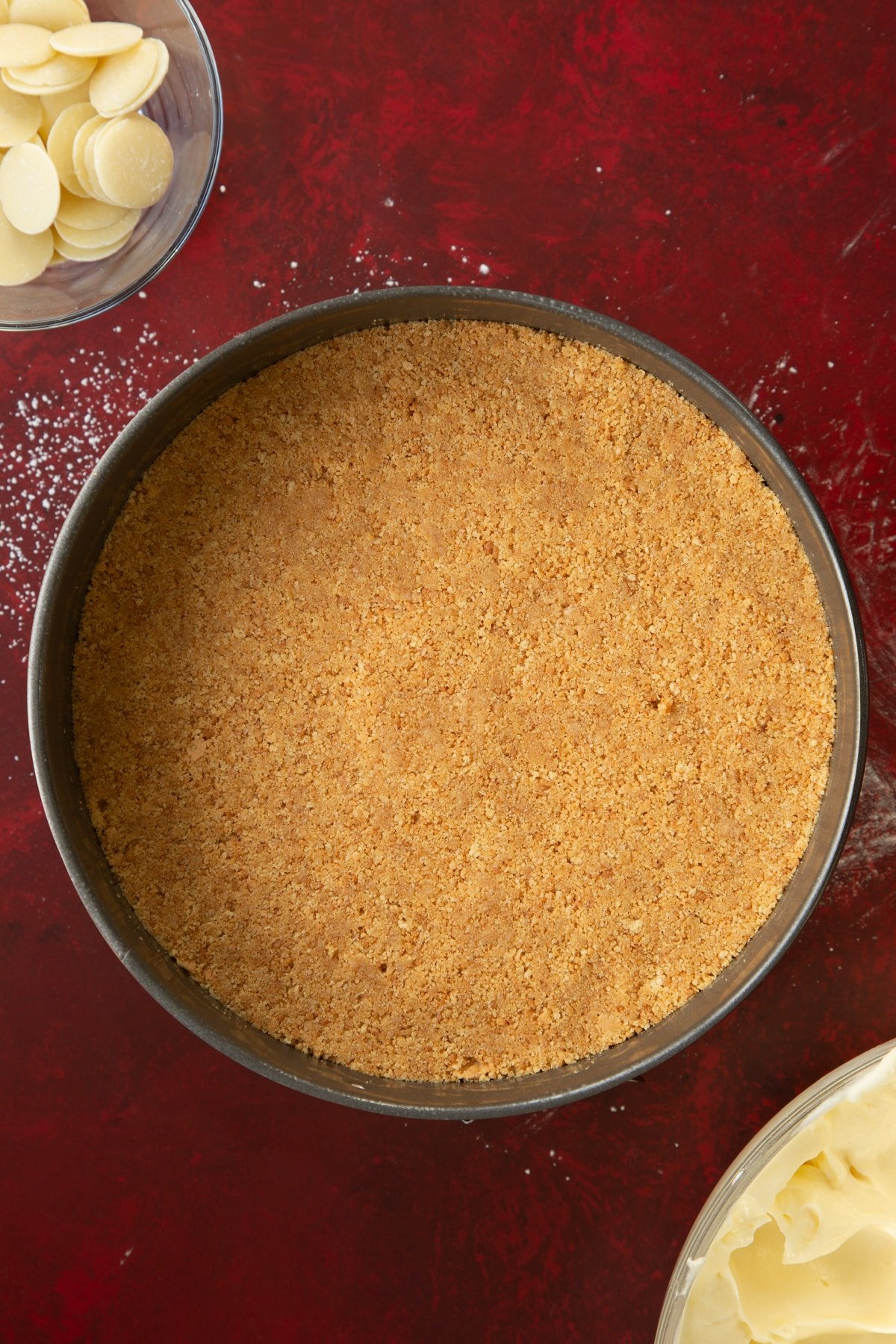 buttery biscuit base pressed down into a round cake tin on a red surface.