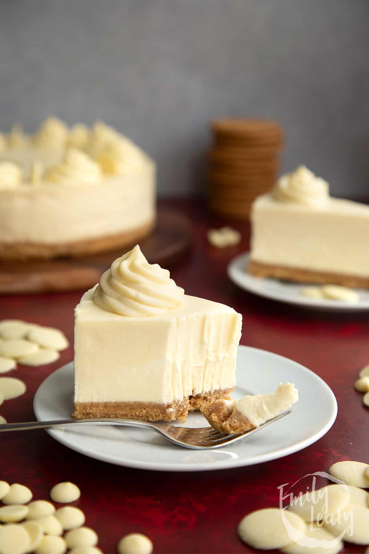 a slice of white chocolate cheesecake on a white plate with a fork breaking off a chunk at the front.
