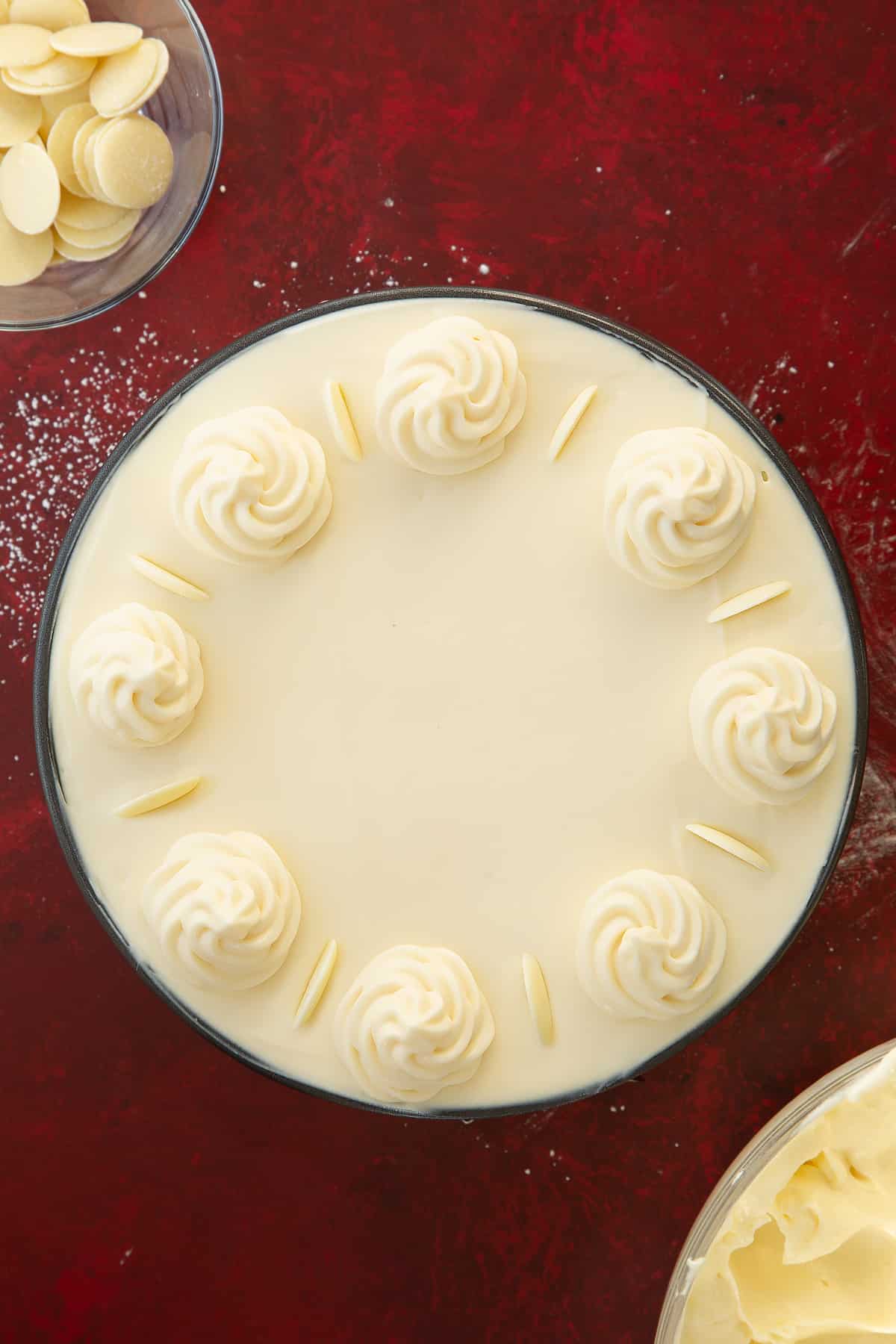 white chocolate cheesecake mixture in a round cake tin filled to the top decorated with white chocolate cheesecake rosettes and white chocolate buttons.
