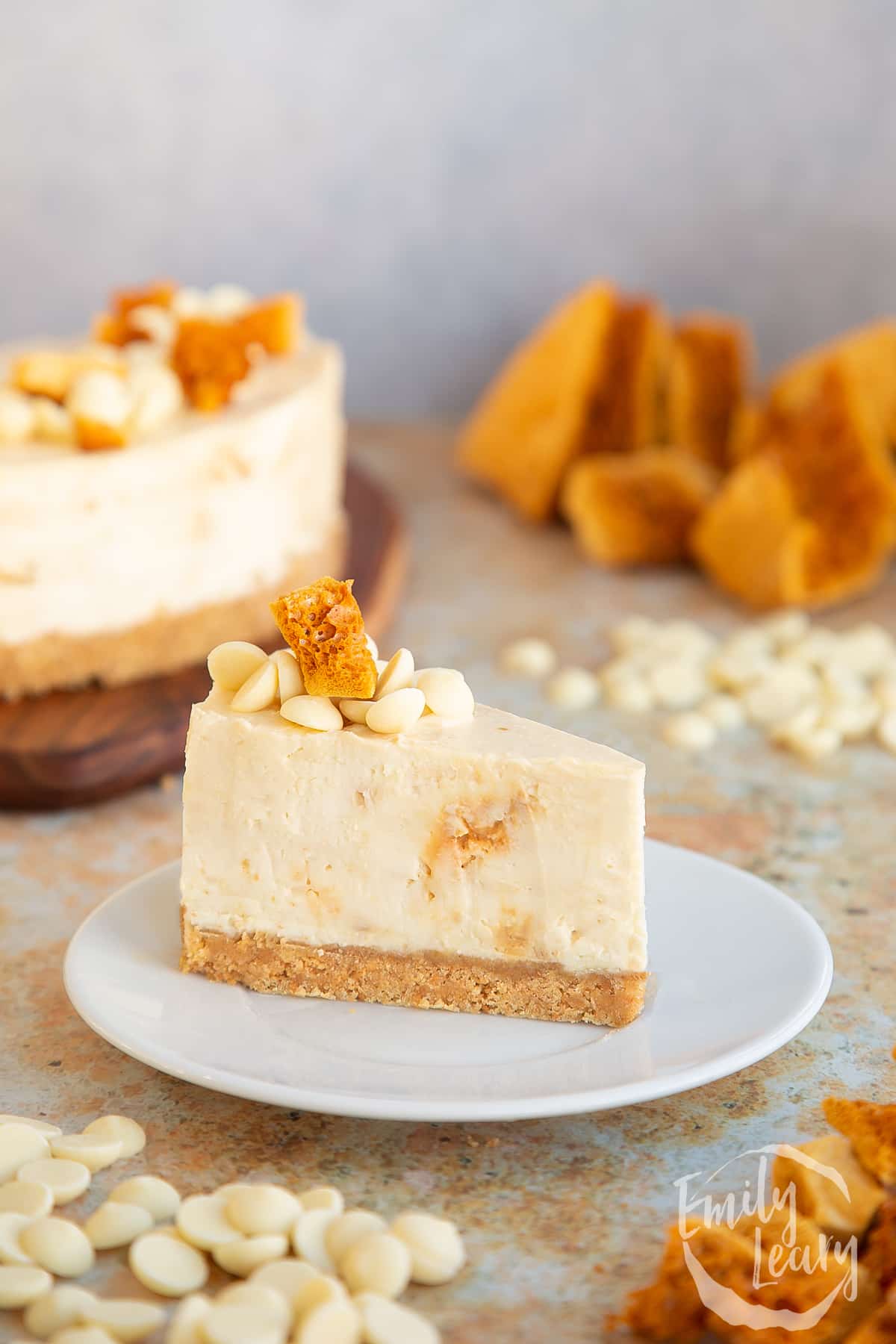 a slice of white chocolate honeycomb cheesecake on a white plate decorated with honeycomb chunks and white chocolate chips.