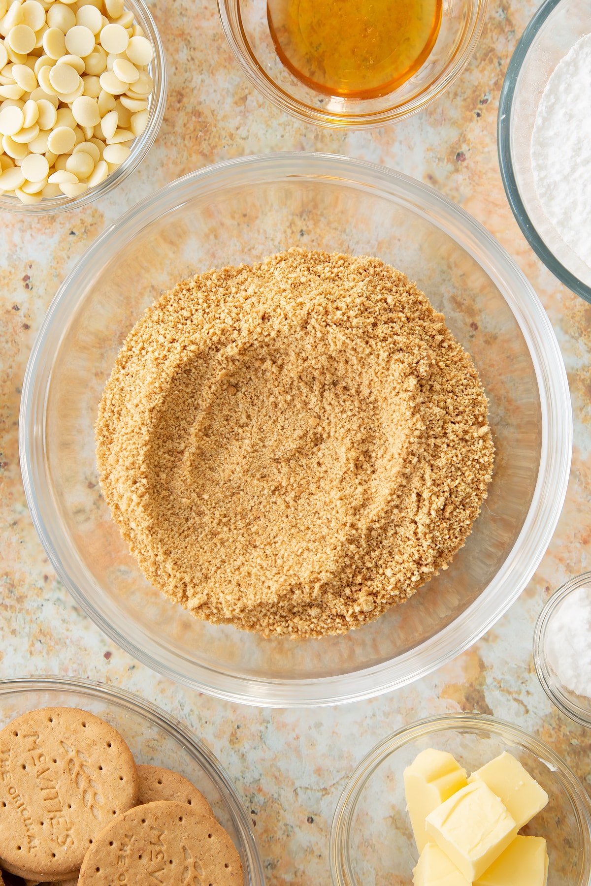 crushed biscuit crumbs in a largw clear bowl surrounded by ingredients in smaller clar bowls.