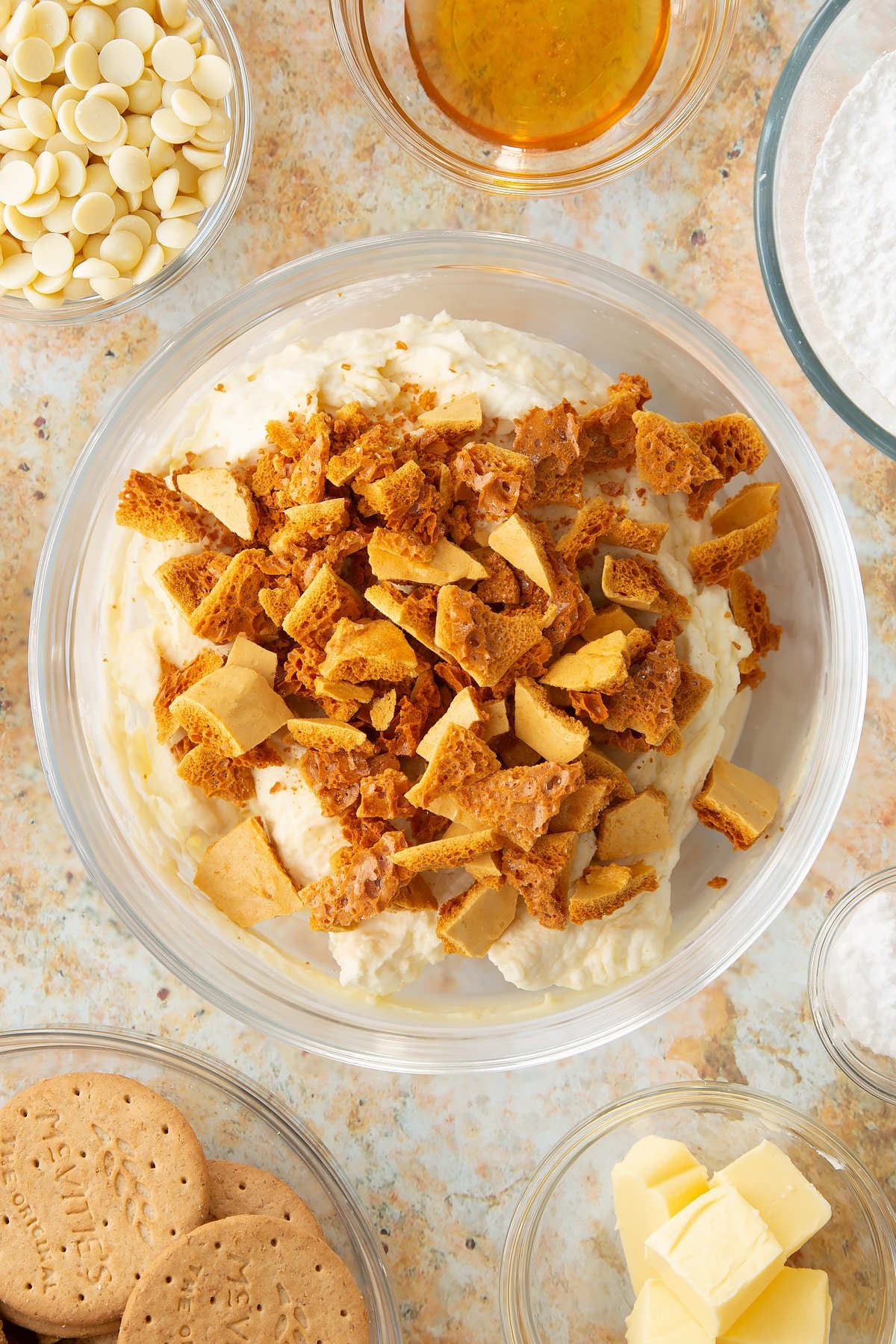 whisked white chocolate cheesecake topping in a large clear bowl topped with honeycomb crumbs,