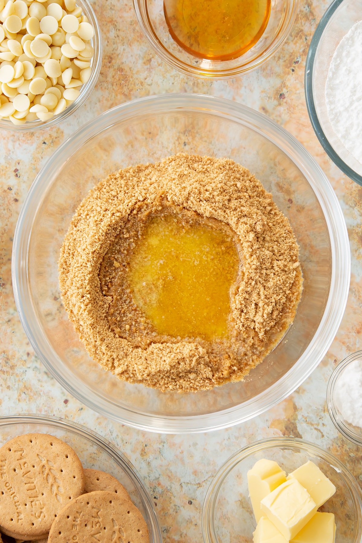 crushed biscuit crumbs topped with melted butter in a large clear bowl surrounded by ingredients in smaller clear bowls.