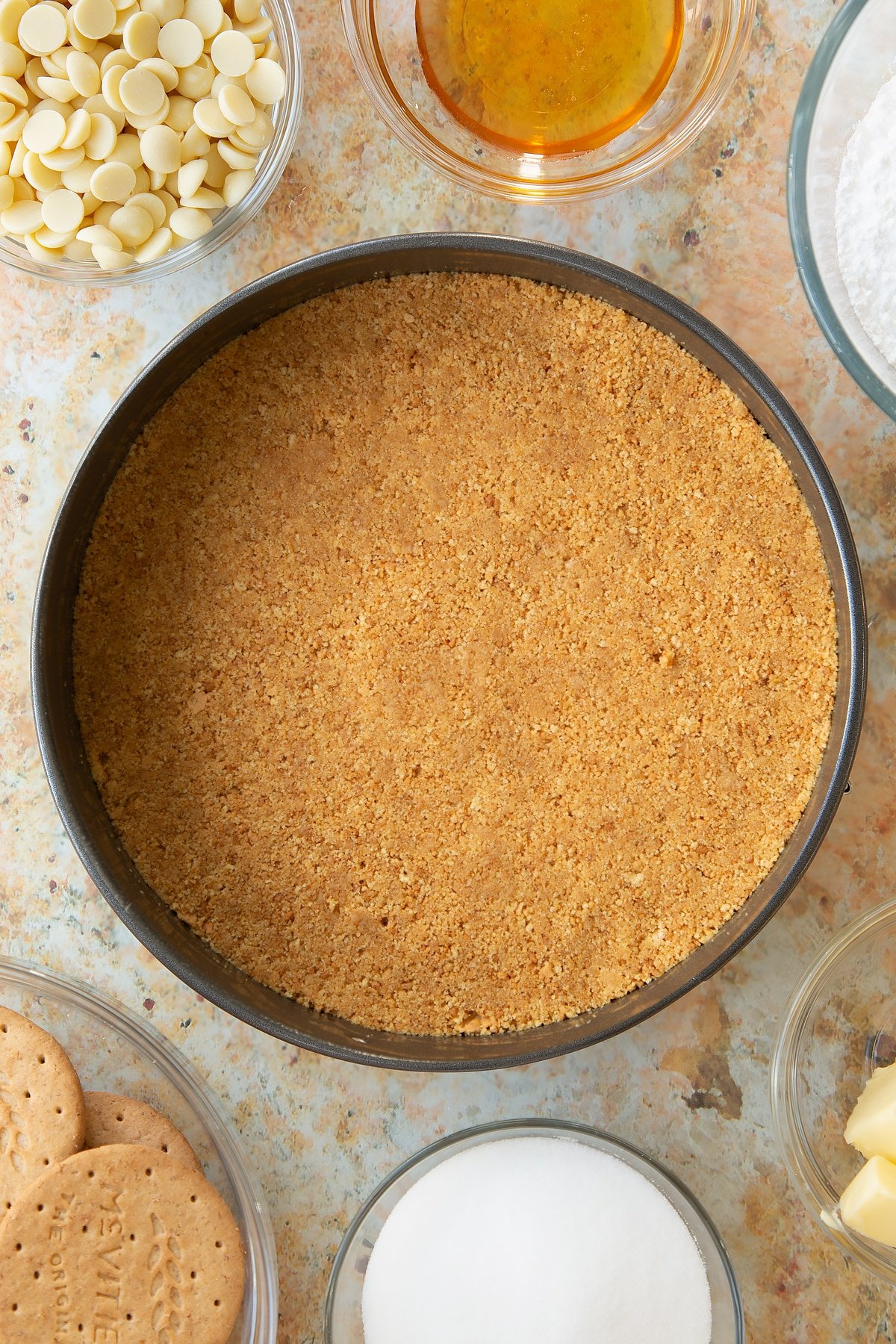 buttery biscuit base pressed down into a metal cake tin surrounded by bowls of ingredients.