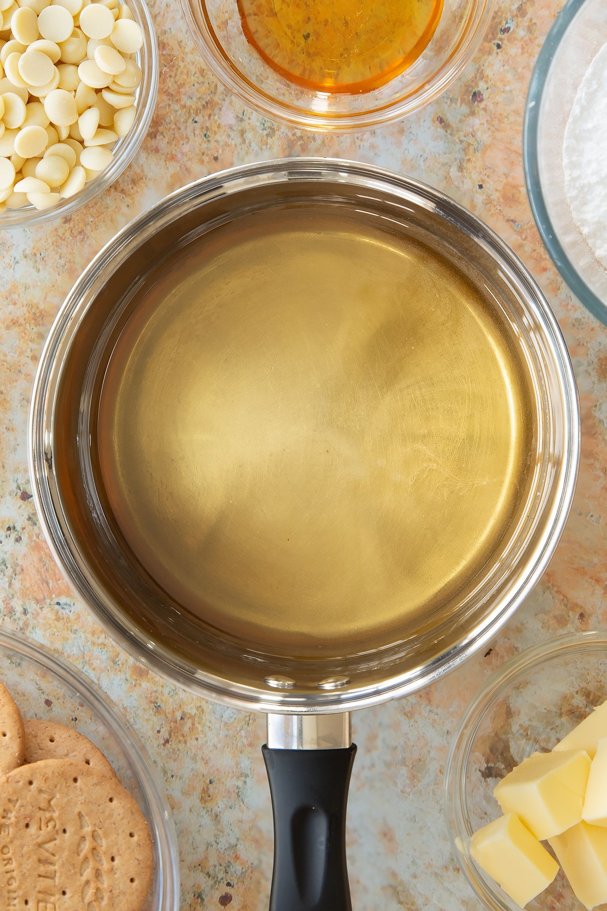 transparent sugar and water mixture in a saucepan surrounded by ingredients in small clear bowl.