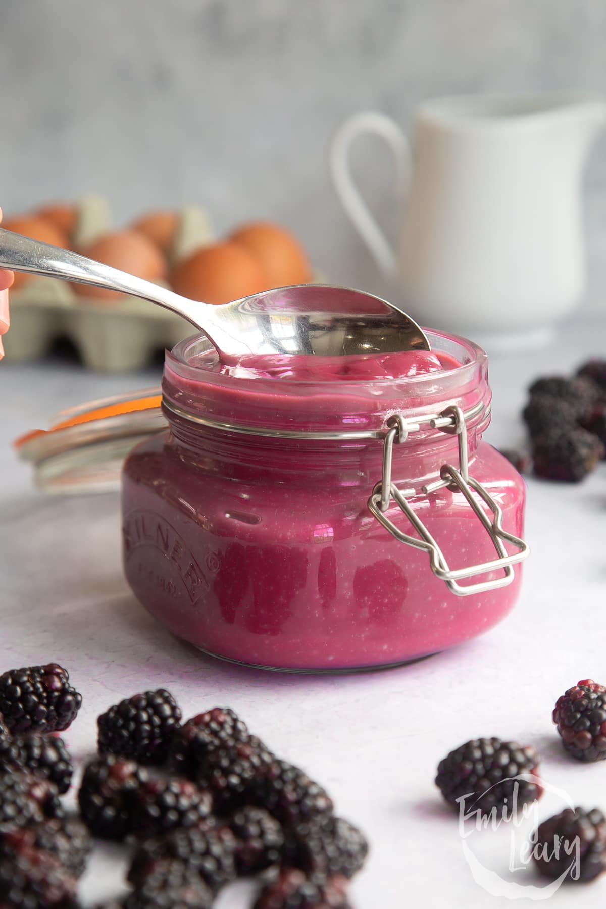 Blackberry curd in a glass jar surrounded by blackberries and a spoon at the top of the jar.