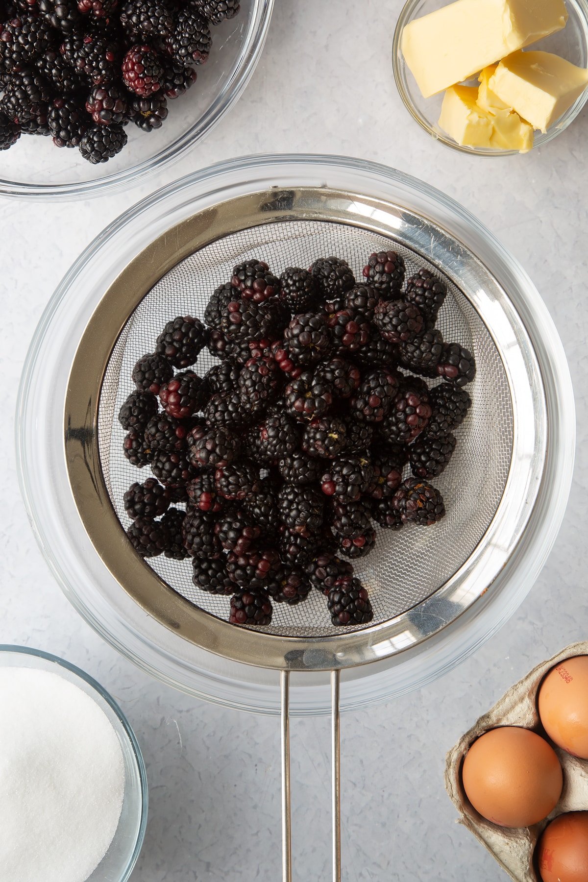 a silver sieve holding blackberries over a large clear bowl.