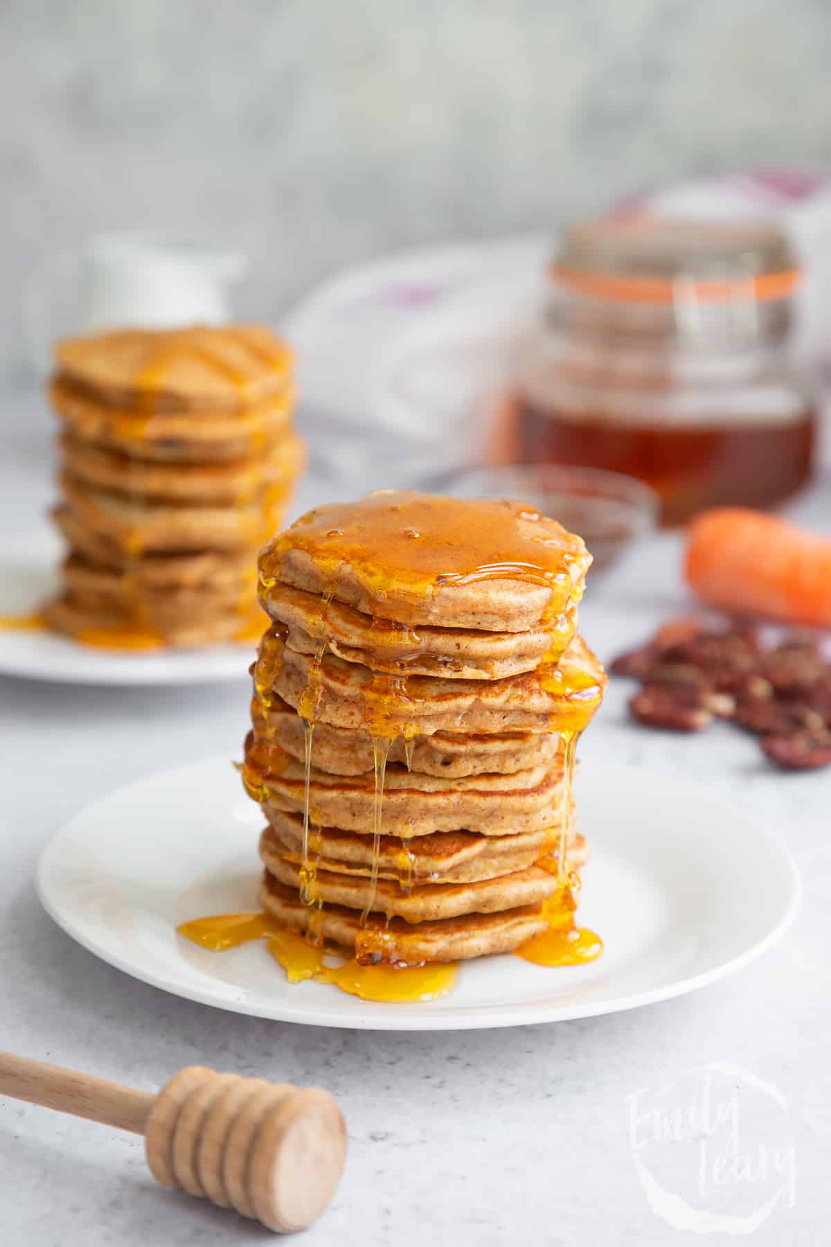 a stack of carrot cake pancakes on a white plare with more pancakes in the background topped with honey.