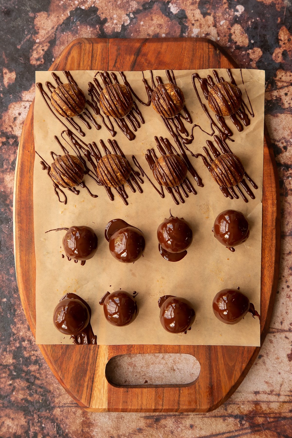 16 small round date energy balls on a parchment lined wooden chopping board 8 drizzled in chocolate and 8 covered in chocolate.