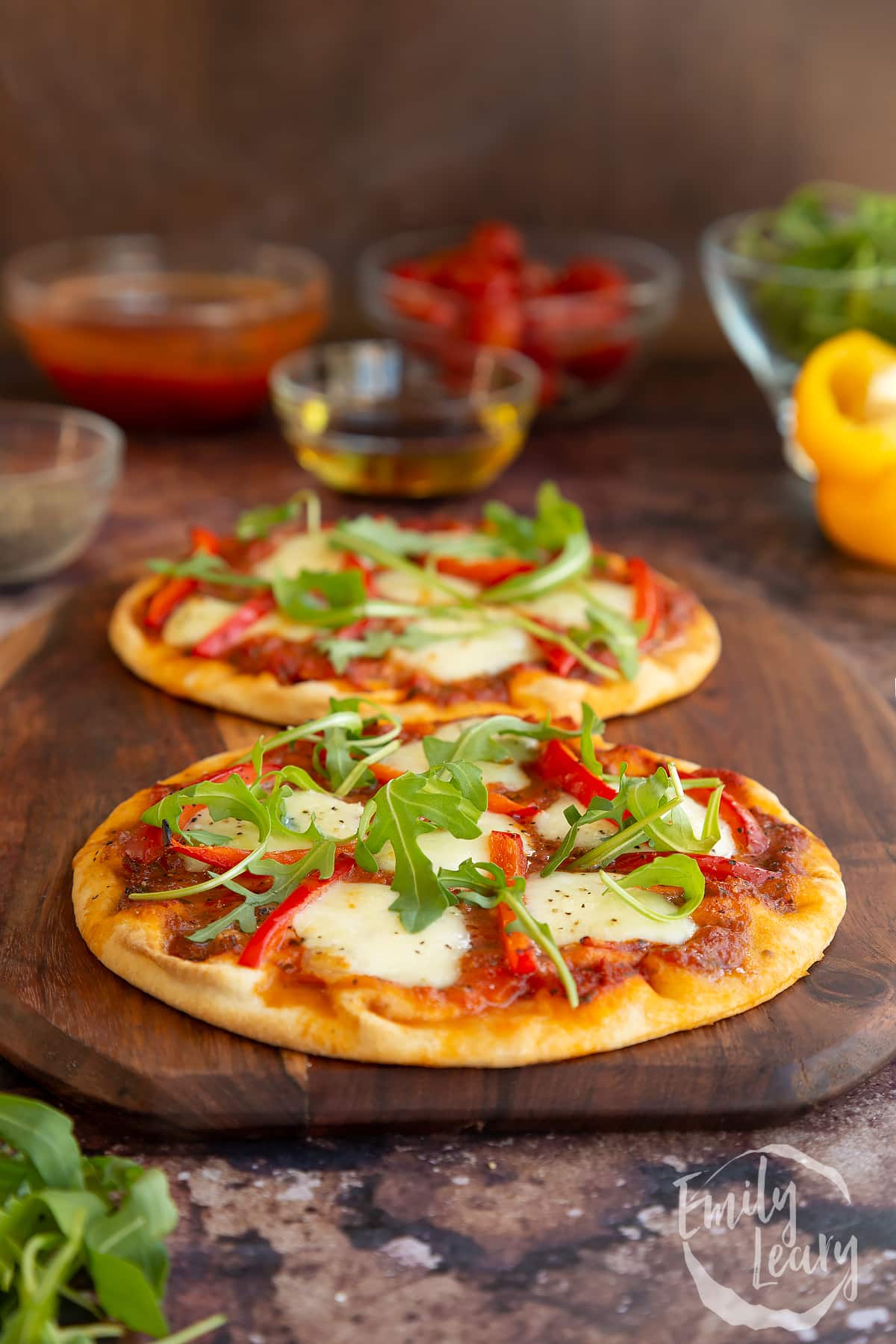 2 Flat bread pizzas on a wooden worktop topped with peppers, rocket and mozzerella.