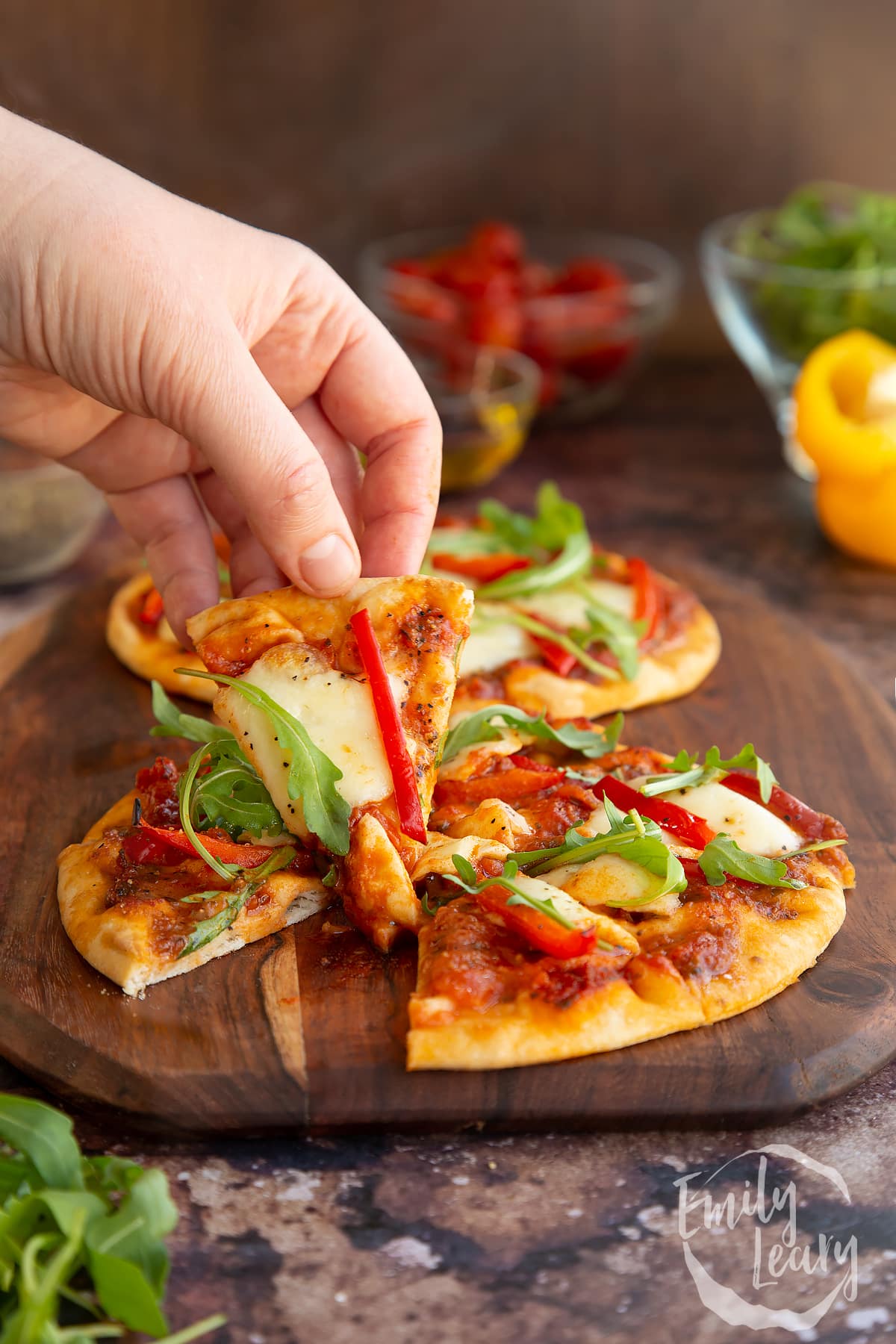 a hand holding a slice of flat bread pizza with the pizza in the background.