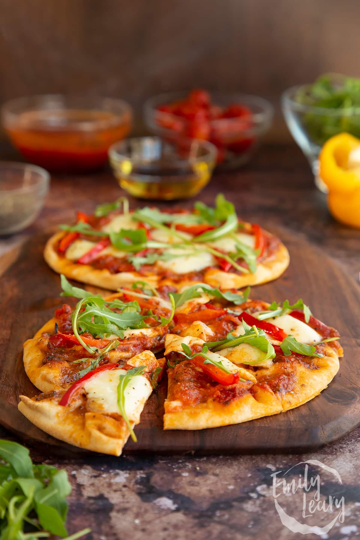 2 baked flat bread pizzas on a wooden chopping board with a slice cut out surrounded by ingredients.