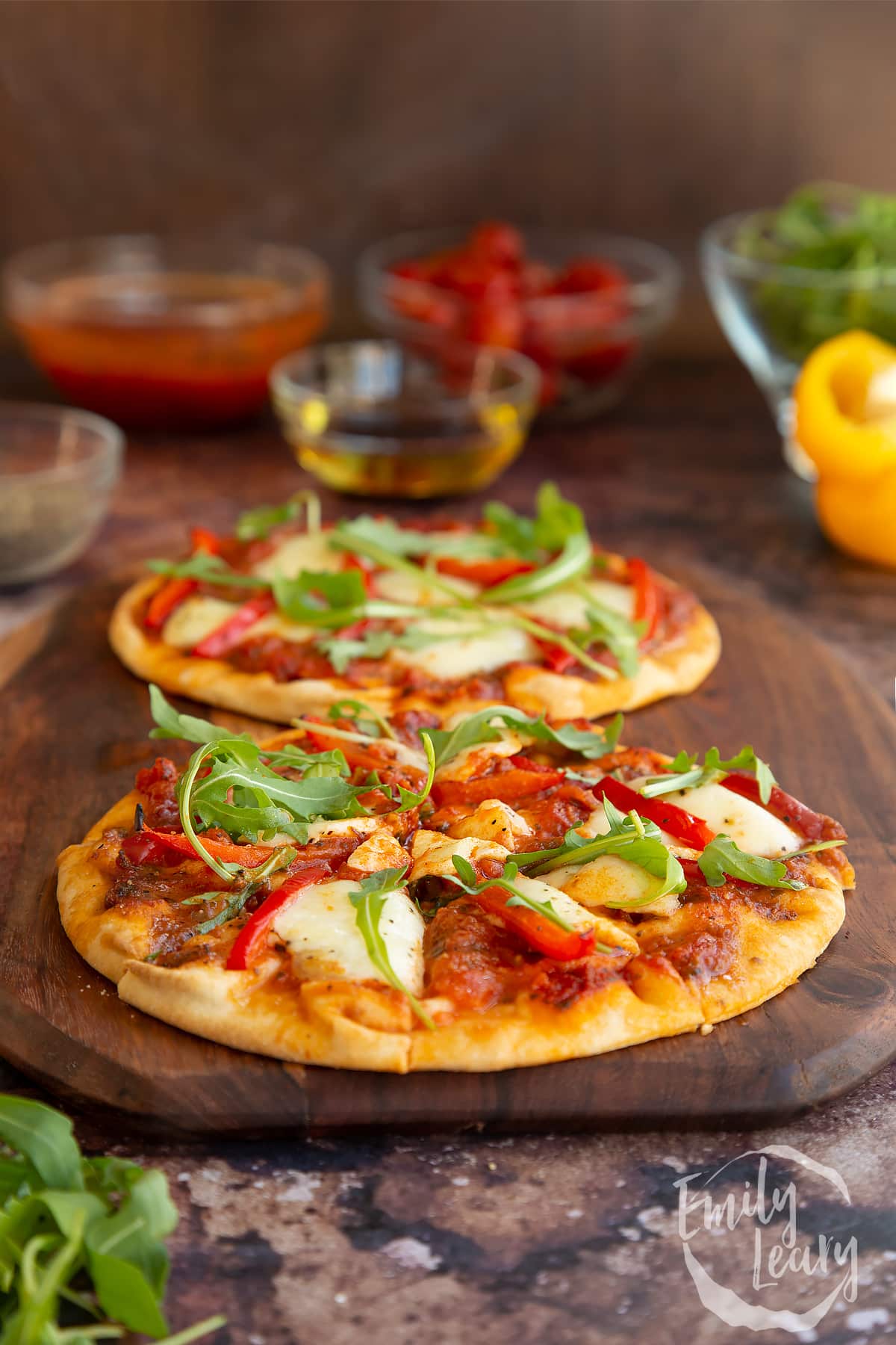 side view of 2 baked flat bread pizzas on a wooden chopping board surrounded by ingredients.