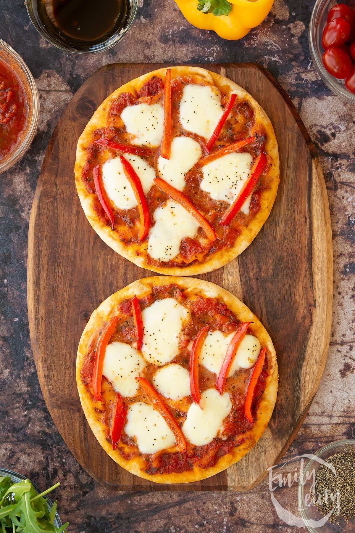 2 baked flat bread pizzas on a wooden chopping board surrounded by ingredients.