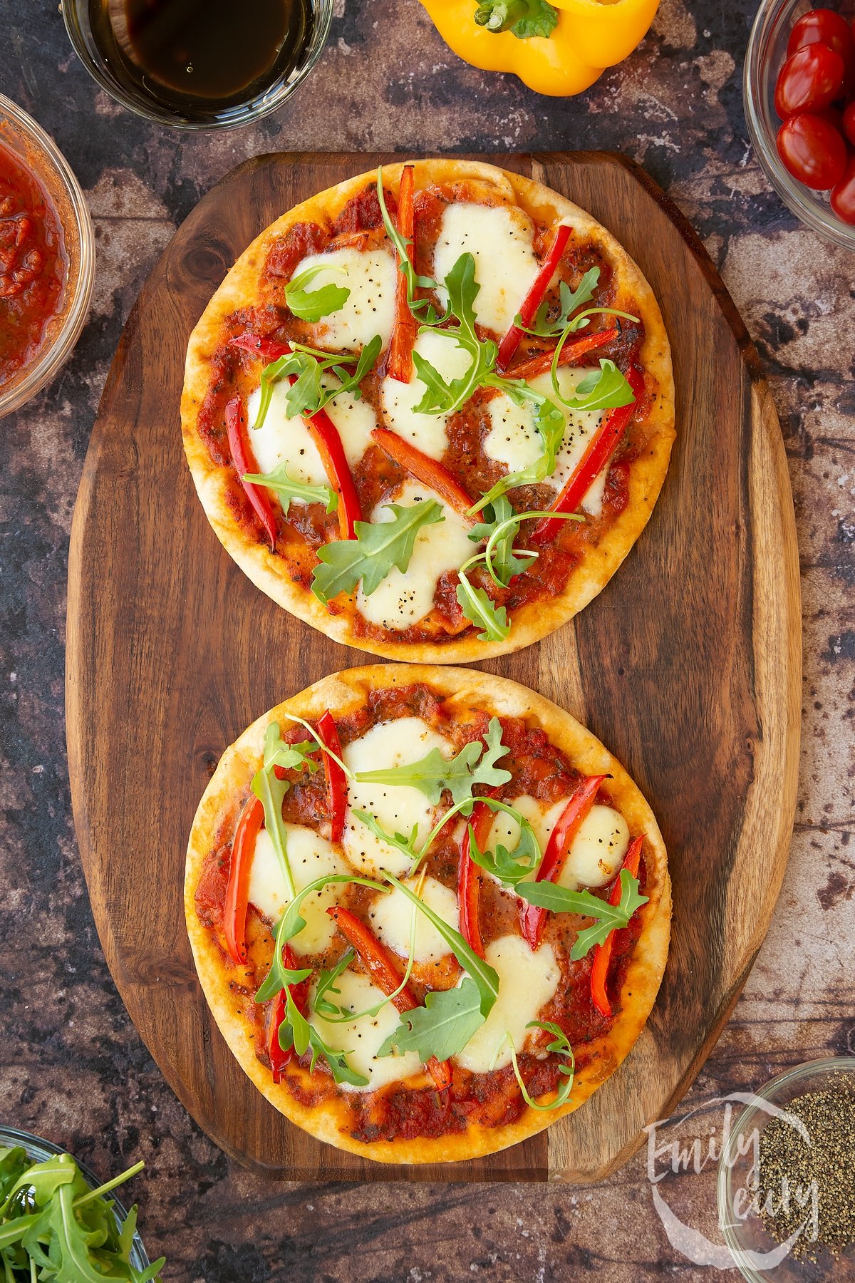 overhead shot of 2 Flat bread pizzas on a wooden worktop topped with peppers, rocket and mozzerella.