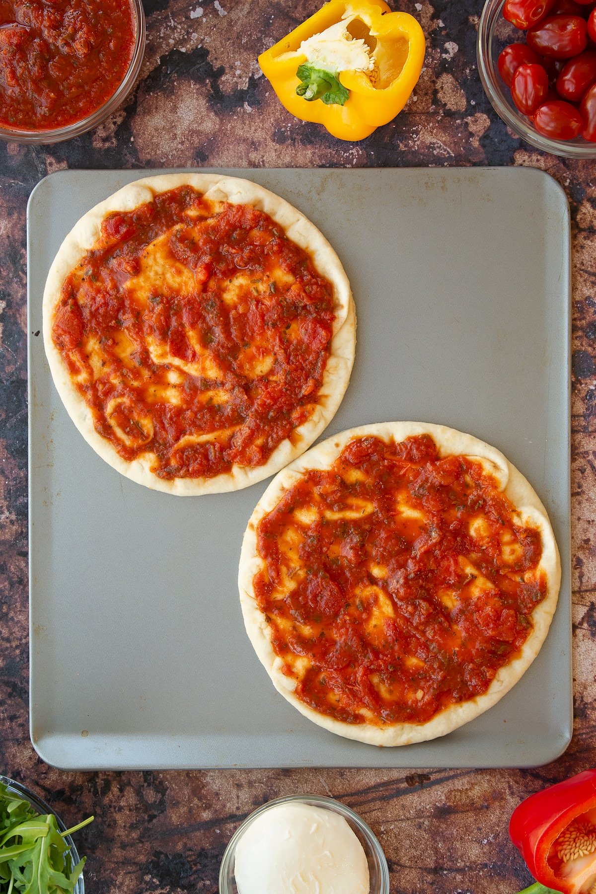 2 flatbreads on a baking tray topped with tomato pasta sauce.