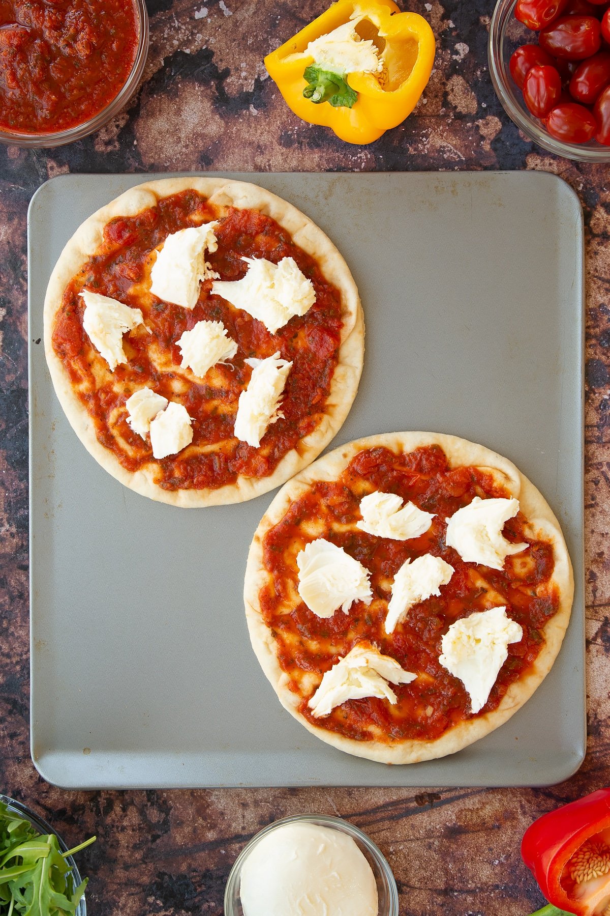 2 flatbreads on a baking tray topped with tomato pasta sauce and pieces of mozzarella.