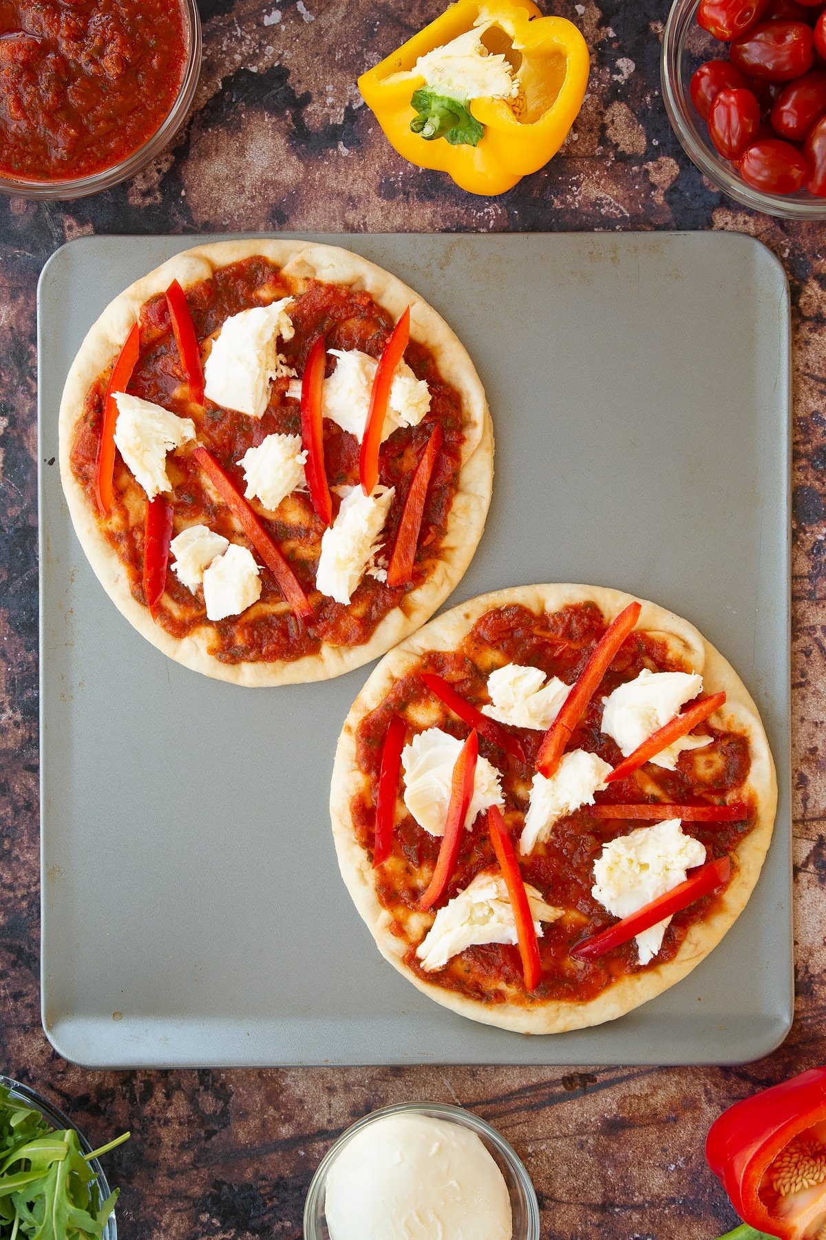 2 flatbreads on a baking tray topped with tomato pasta sauce, pieces of mozzarella and red sliced peppers.
