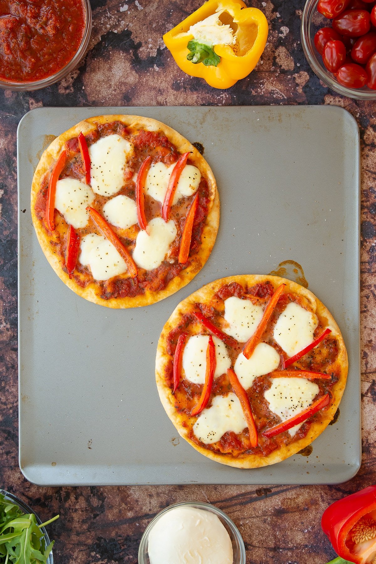 2 baked flatbread topped with peppers and mozzarella drizzled with oil and pepper on a large baking tray.