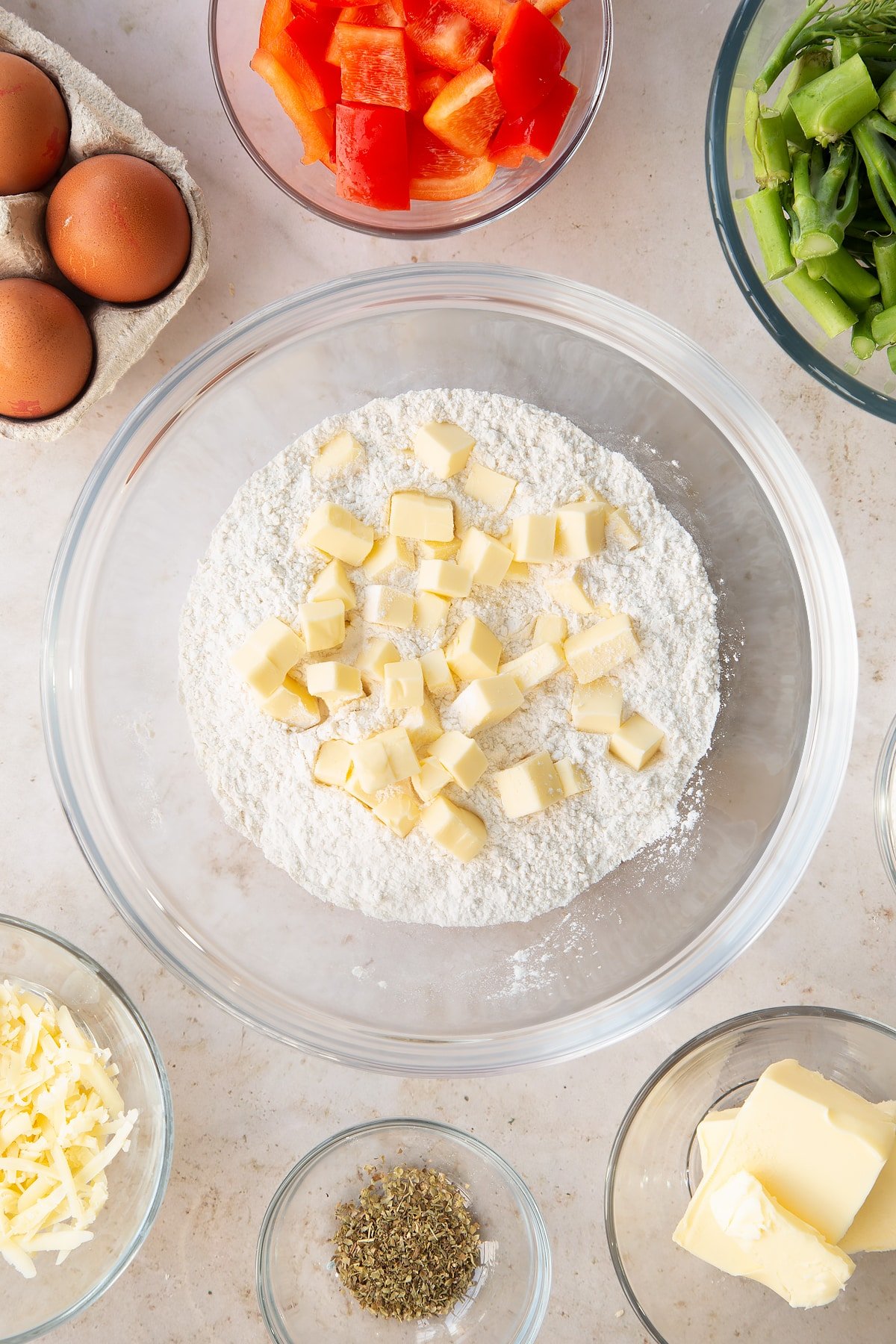 Flour and cubed butter in a large clear mixing bowl surrounded by ingredients.