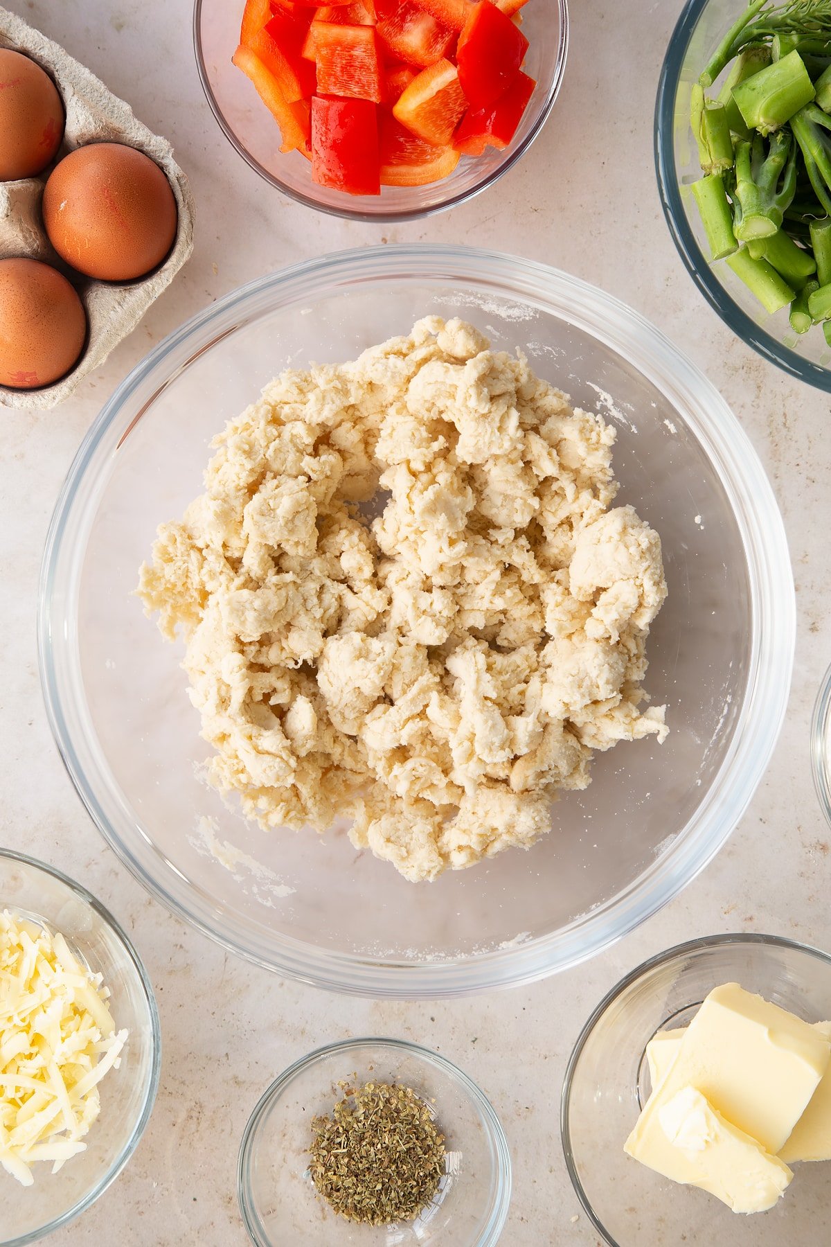 shortcrust pastry dough crumbled in a large clear bowl surrounded by ingredients.