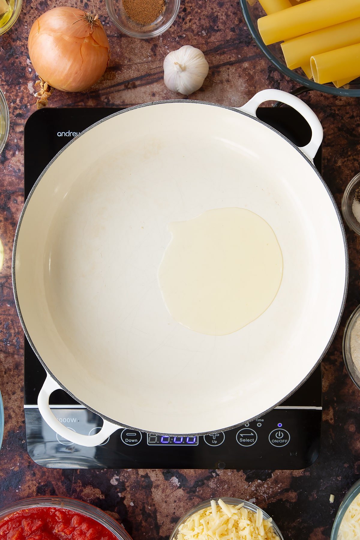 Overhead shot of a pan on an induction hob with oil surrounded by some of the ingredients required to make the Spinach and ricotta cannelloni.