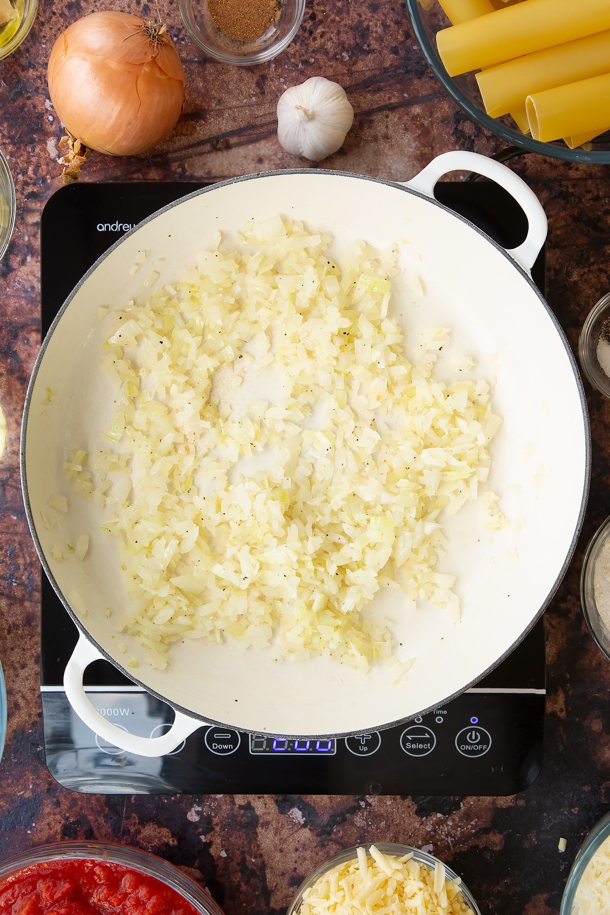 Chopped onion and garlic inside a pan on a induction hob surrounded by some of the other ingredients required to make the recipe.