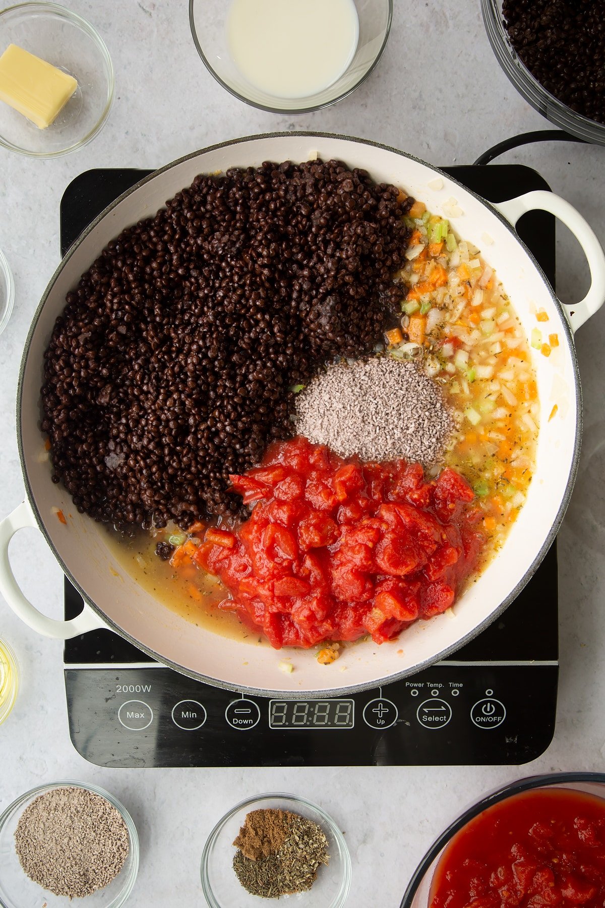 a large frying pan on an induction hob with mixed vegetables, brown lentils and tinned tomatos.